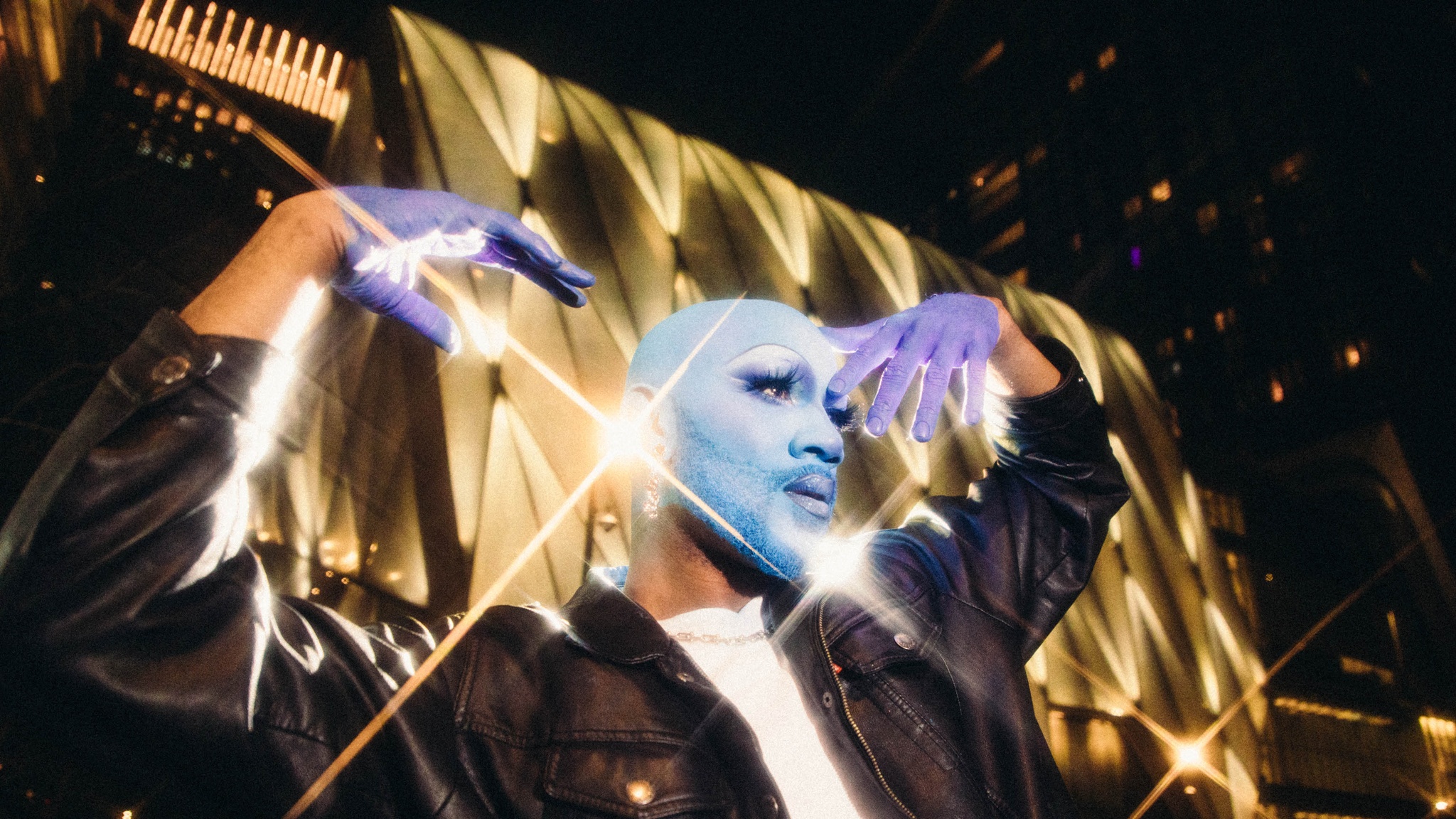 Cain Coleman, a Black multihyphenate drag performer, poses at night outside The Shed with their head and face covered in blue makeup. Cain wears a black leather jacket and white t-shirt, holding their hands, covered in purple makeup to resemble gloves, up to either side of their face. Behind them, lights flash off the reflective surface of The Shed in small starbursts. 