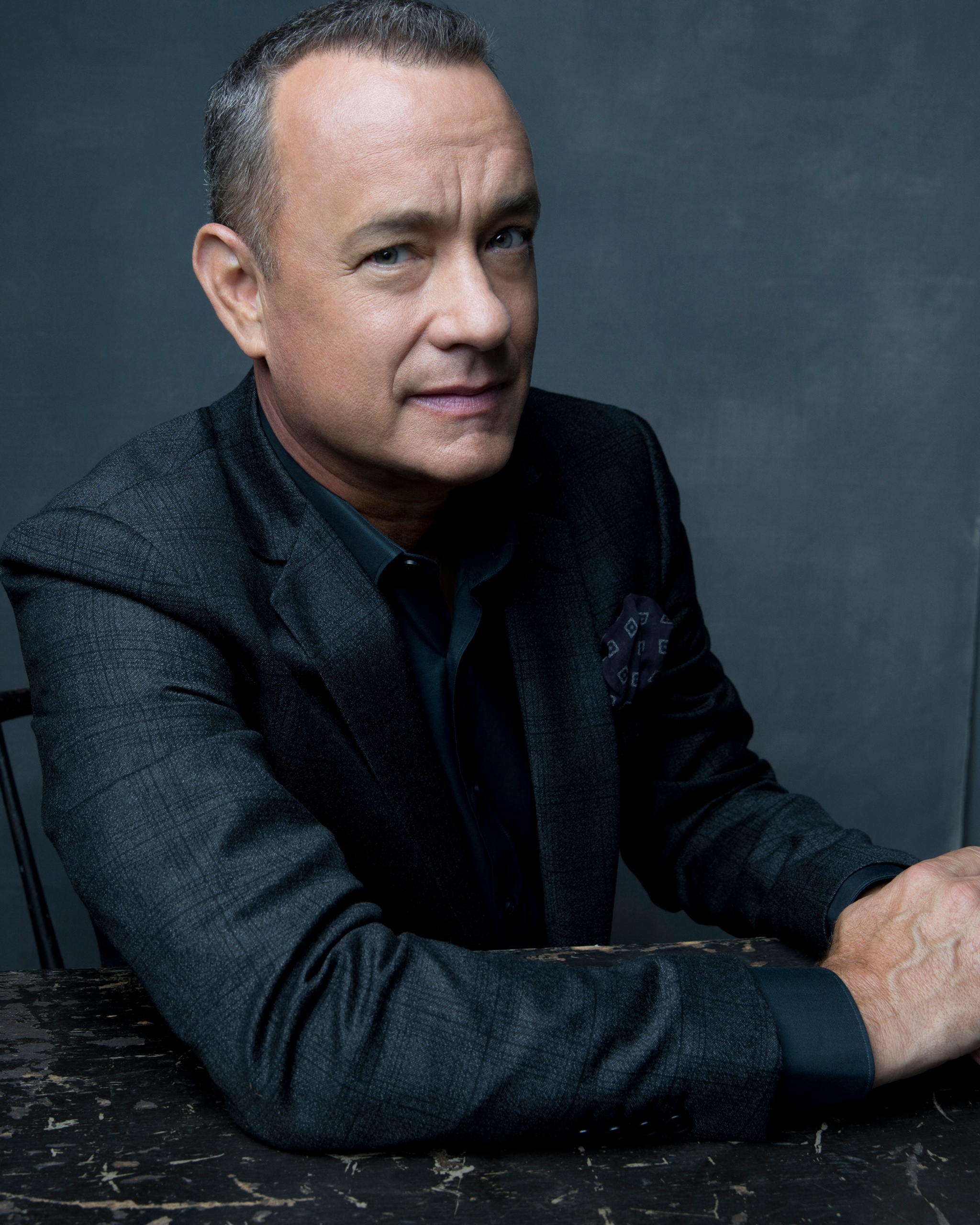 A portrait of actor Tom Hanks. Tom is a white, middle-aged man who wears a charcoal gray suit, sitting at a dark brown wooden table and leaning with one shoulder forward and hands clasped on the tabletop. He looks at us from the side with a knowing smile.