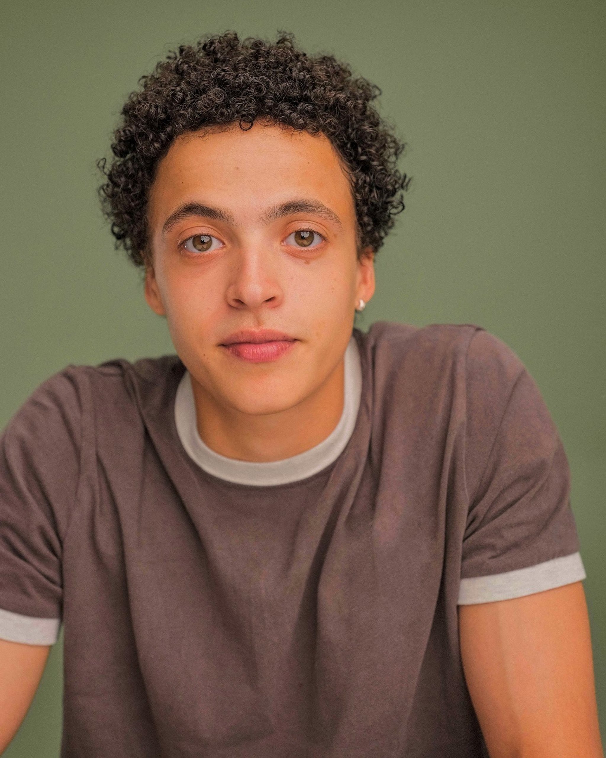 A photo portrait of nonbinary actor Lee Braithwaite. Lee faces us directly and is pictured from the chest up, leaning slightly forward with shoulders arched as they support themselves. They wear a brown t-shirt with gray rings around the neck and sleeves. They look at us with an open, slightly inquisitive look, with right eyebrow raised. Their brown hair is short in tight curls.