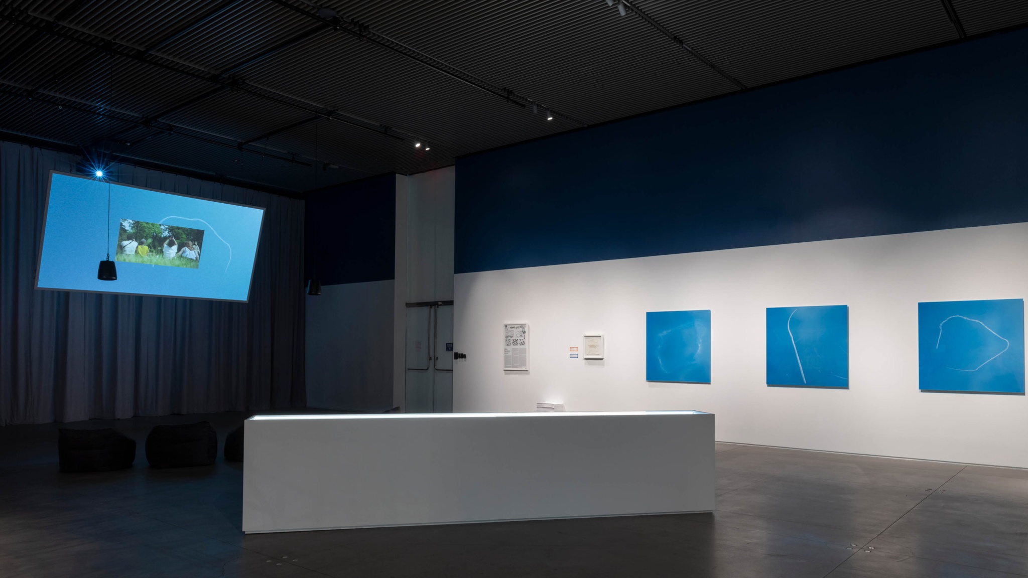 An art installation including a film and photographic prints. The film documents a skywriting performance and is projected on a screen that hangs from the ceiling on a diagonal angle to the left of the space. Along the wall on the right hang three photos in a row of the clouds formed by the skywriting against a bright blue sky. 