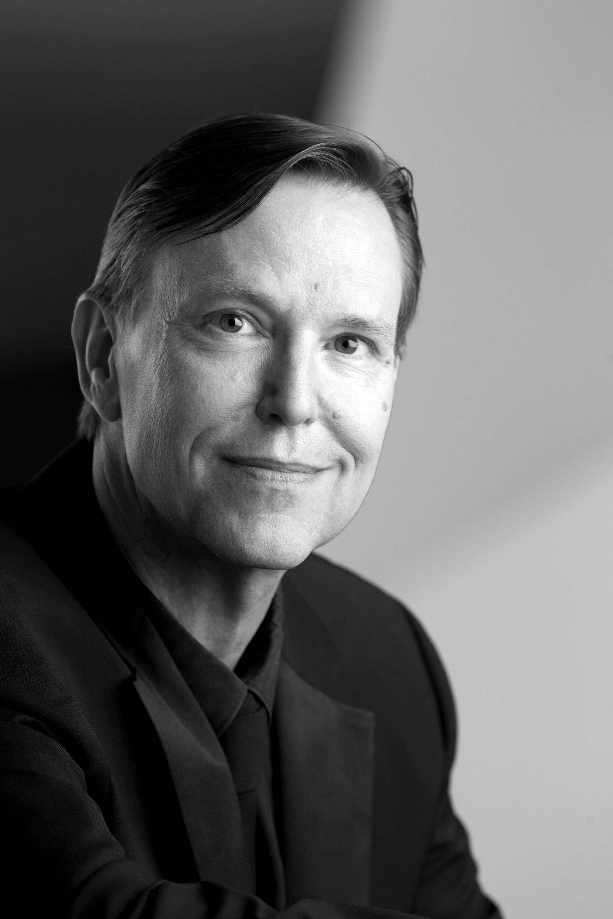 A black and white headshot of conductor Grant Gershon, a white man with brown hair parted to the side with a slight swoop over his forehead. Grant wears a black dinner jacket over a black button-up shirt and smiles while looking directly at us. 