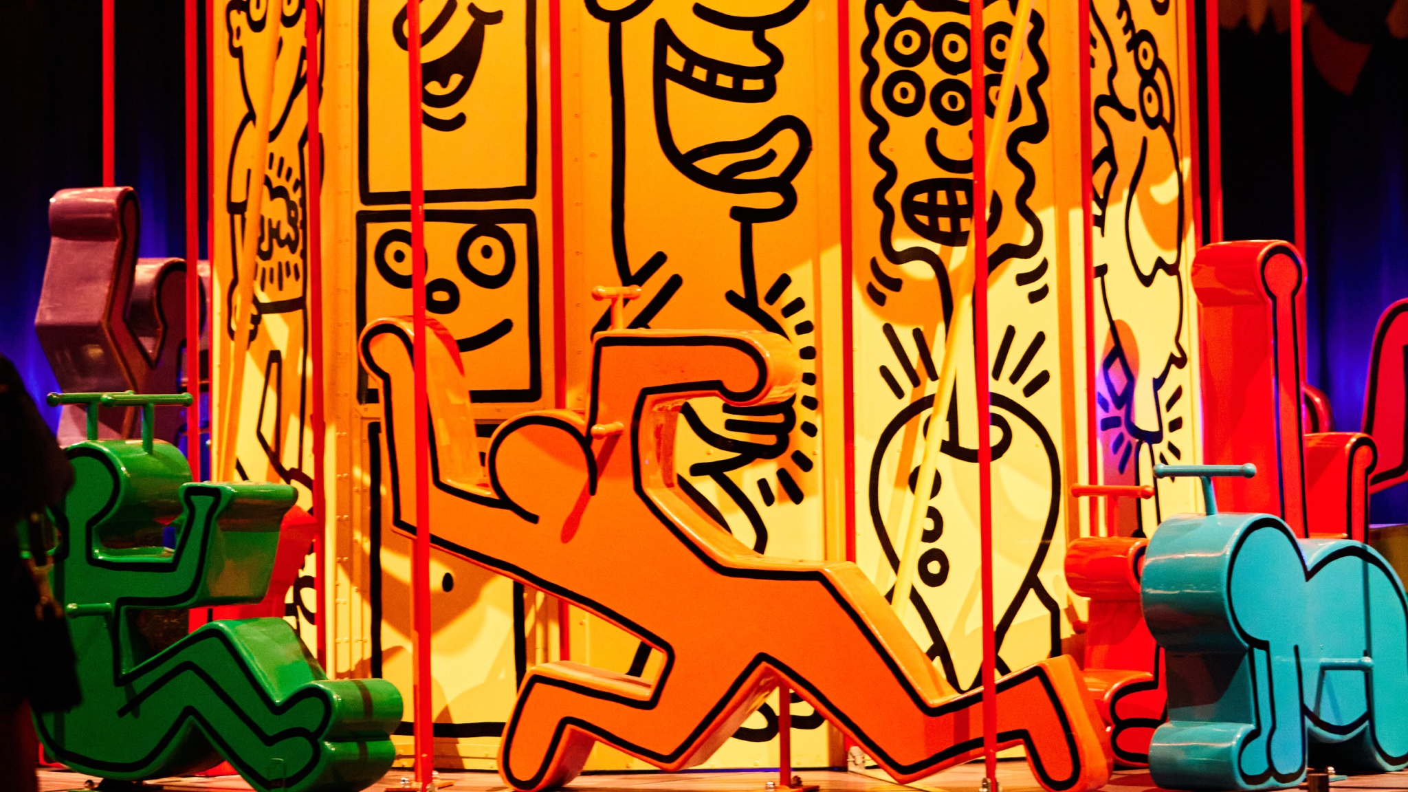 A close up photo of a carousel ride designed by artist Keith Haring. Its yellow interior column is covered in black cartoonish line drawings and sculptures of Haring's distinctive cartoon designs of people are lined around the edge of the ride. 