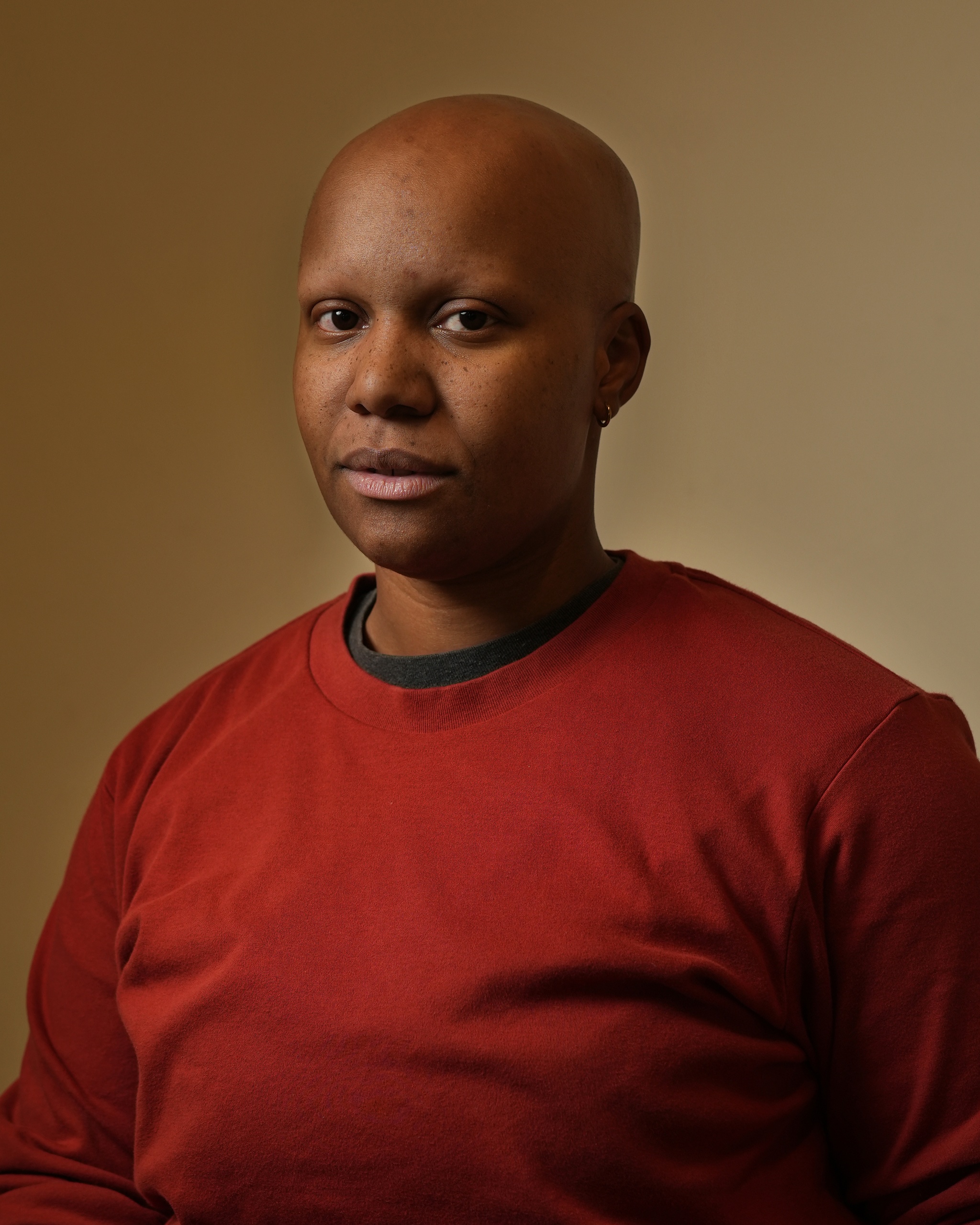 A portrait of Ash Winkfield against a neutral brown background. Ash has a clean shaven head and wears a red longsleeved tshirt. They look directly at us. 