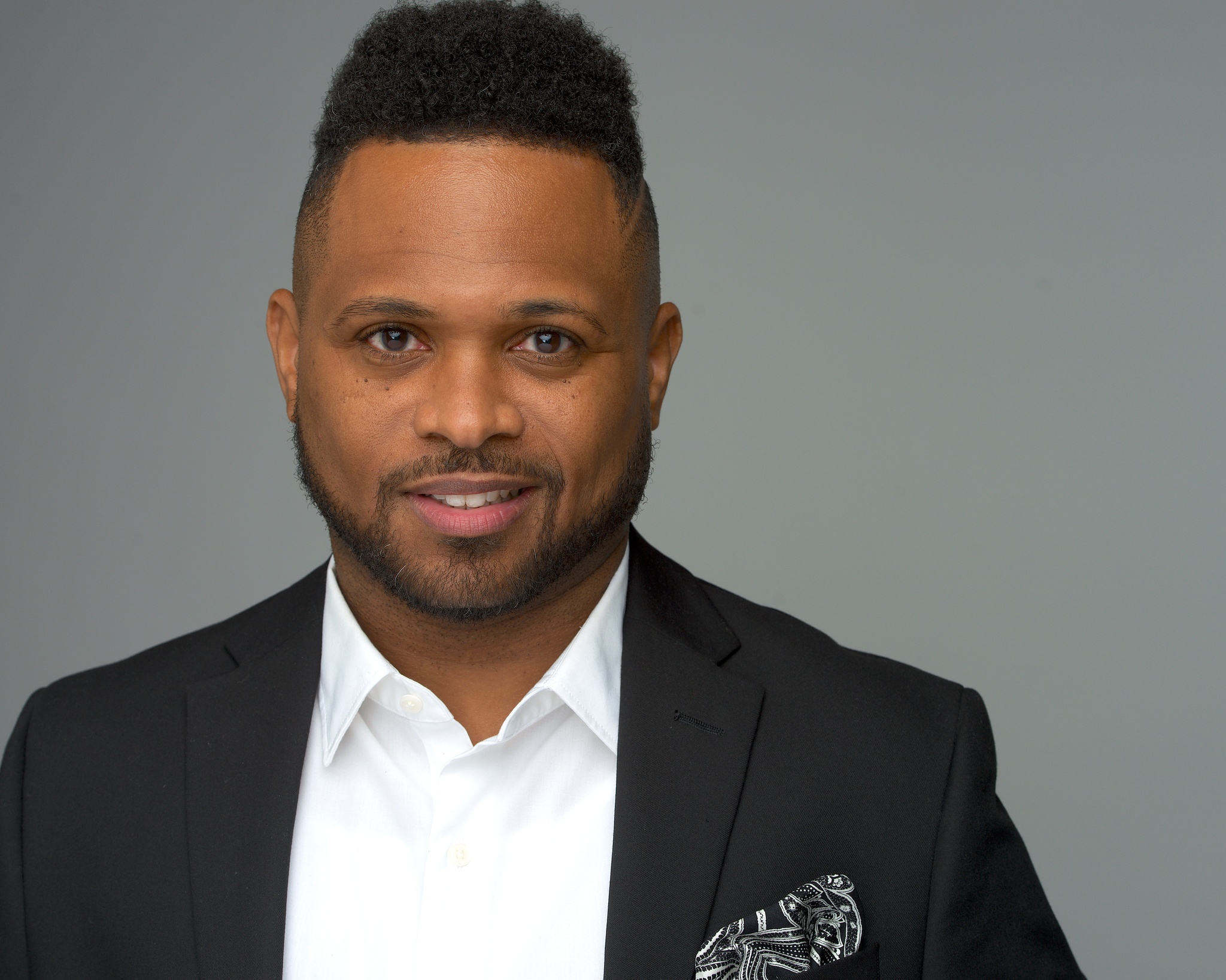 A headshot of Aaron L. McKinney, a Black man who poses facing us directly. He has a well manicured beard and thin mustache and wears a suit blazer with a pocket square. Photo by David Genik