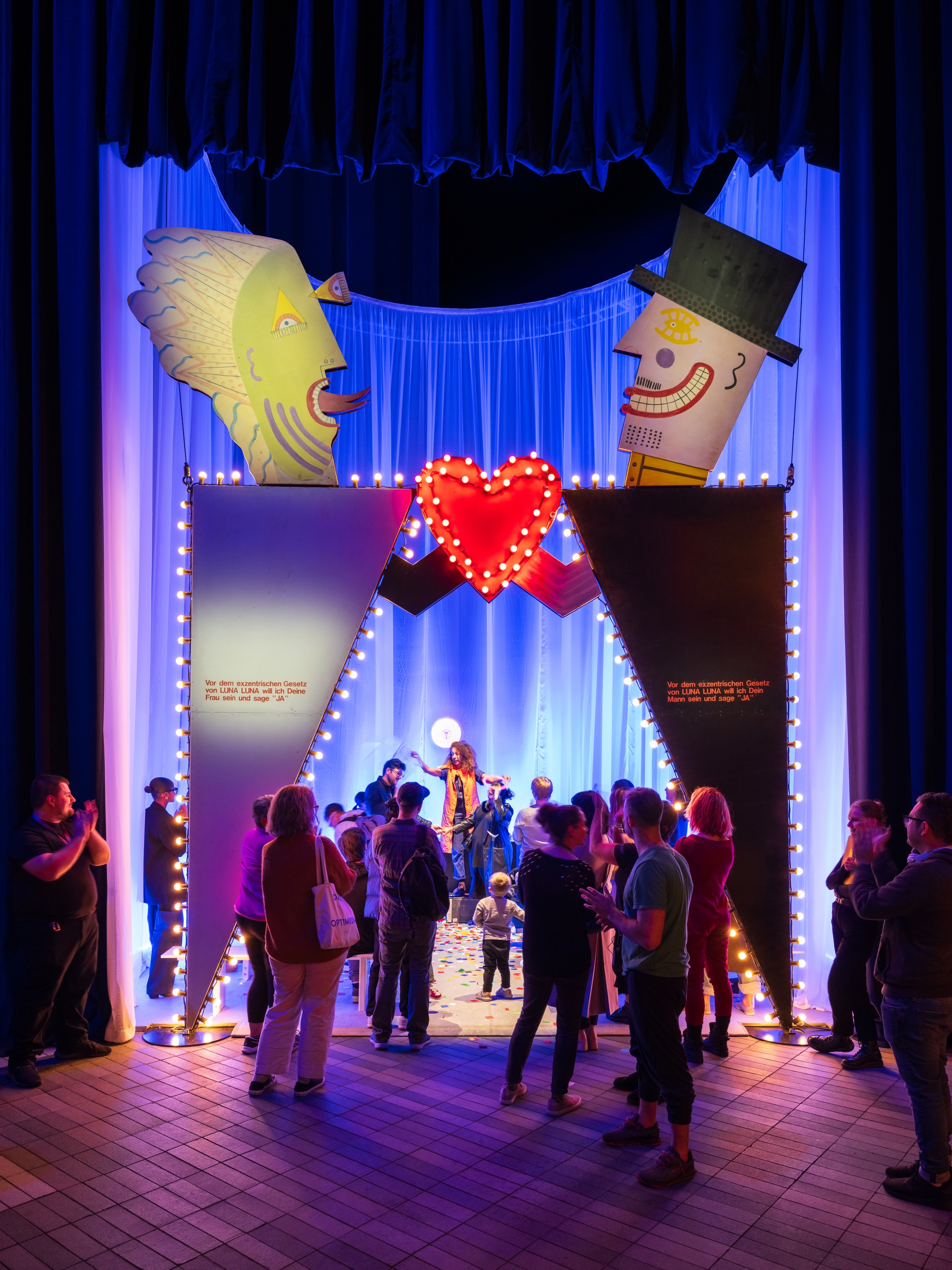 A group of gallery visitors crowd around an art wedding chapel to watch the ceremony being performed inside. The chapel has an entryway that includes stylized triangles resembling bodies. Atop them are cartoonish heads, and the two figures hold a heart lit by electric lights above the center of the entrance.