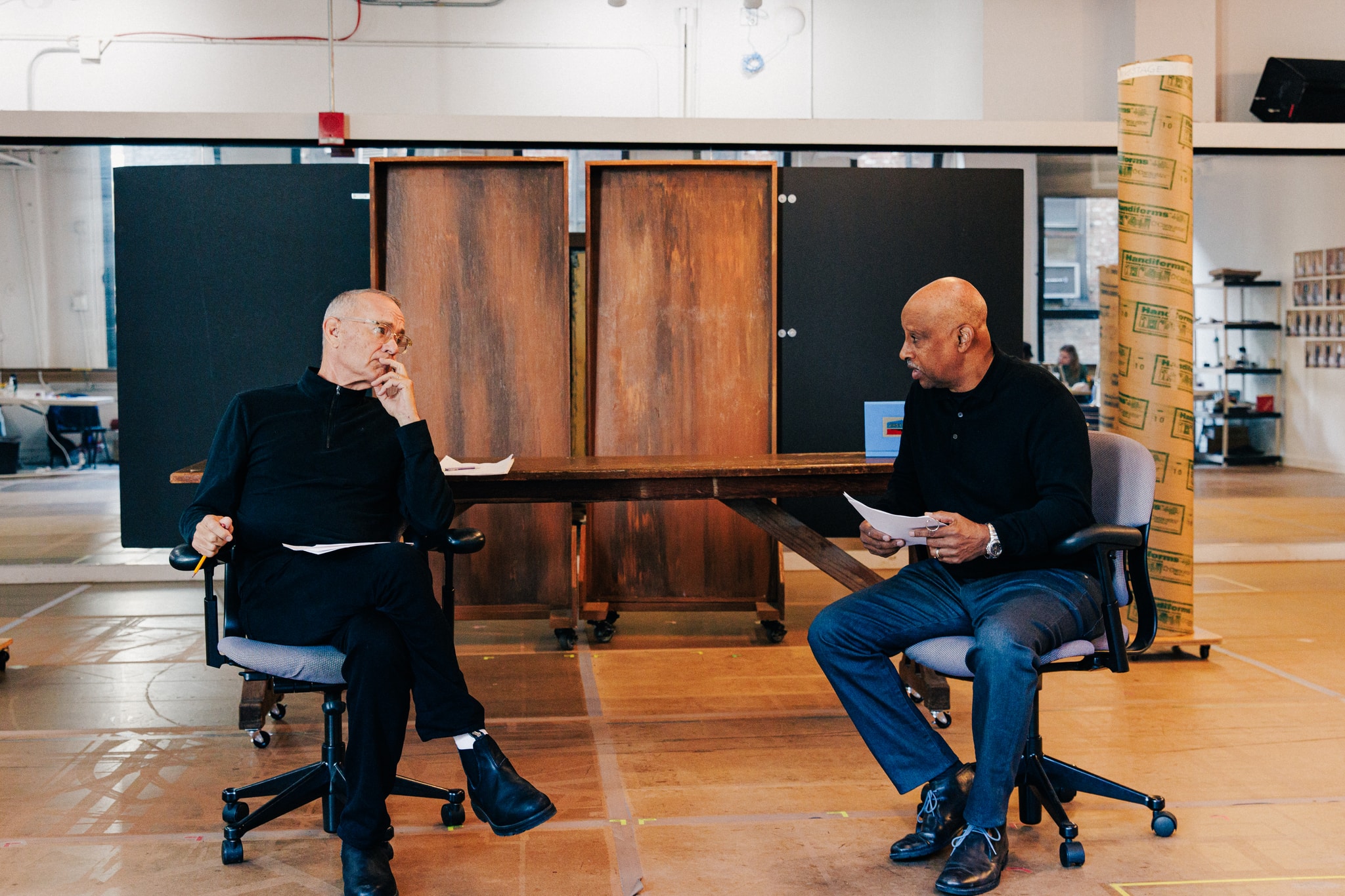 Actors Tom Hanks, a middle aged white man, and Ruben Santiago-Hudson, a middle aged Black man with a clean shaven head, sit in a rehearsal studio in rolling desk chairs facing each other in the middle of a conversation.