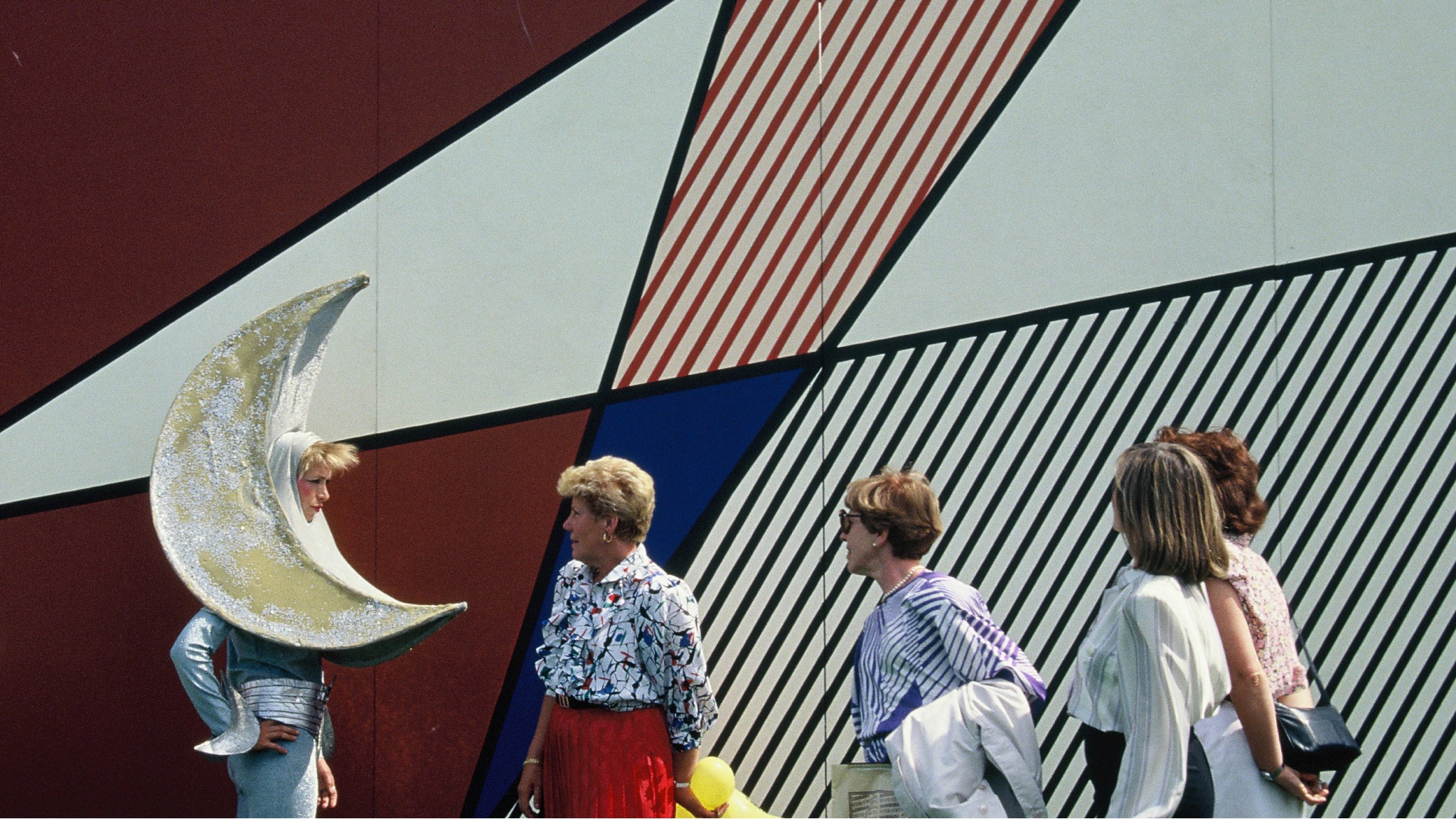 Visitors to the original 1987 Luna Luna carnival interact with a person dressed in a costume with a half moon for a head. The four visitors are lined up in front of a mural by artist Roy Lichtenstein, with red, white, and blue shapes made by diagonal lines. 