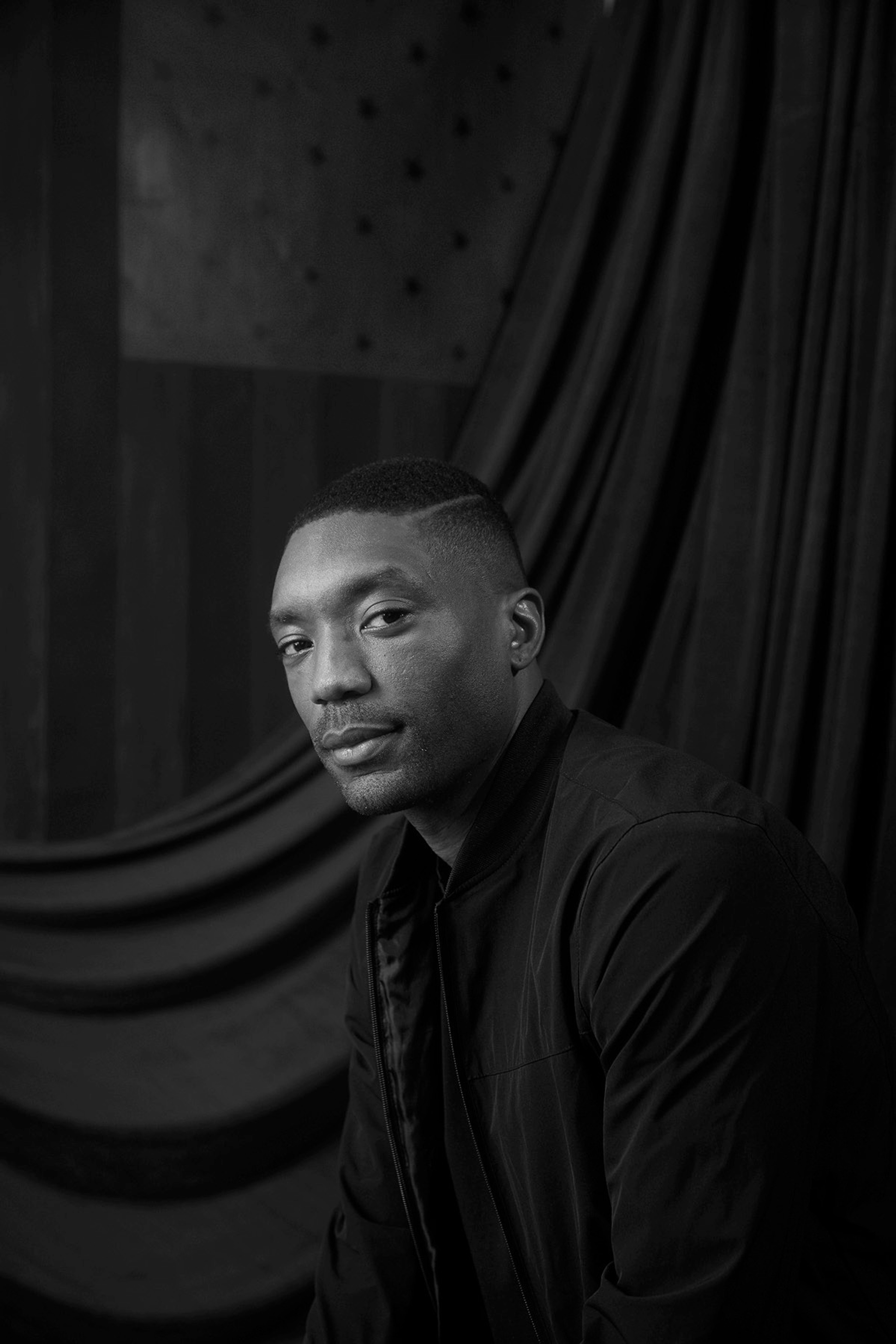 A black-and-white photo of Cyrus Aaron. Aaron's body is in three-quarters view and he turns his head to look at the camera with a slight smile. In the background are black swaths of fabric and the outline of a flag in tones of gray and black. 