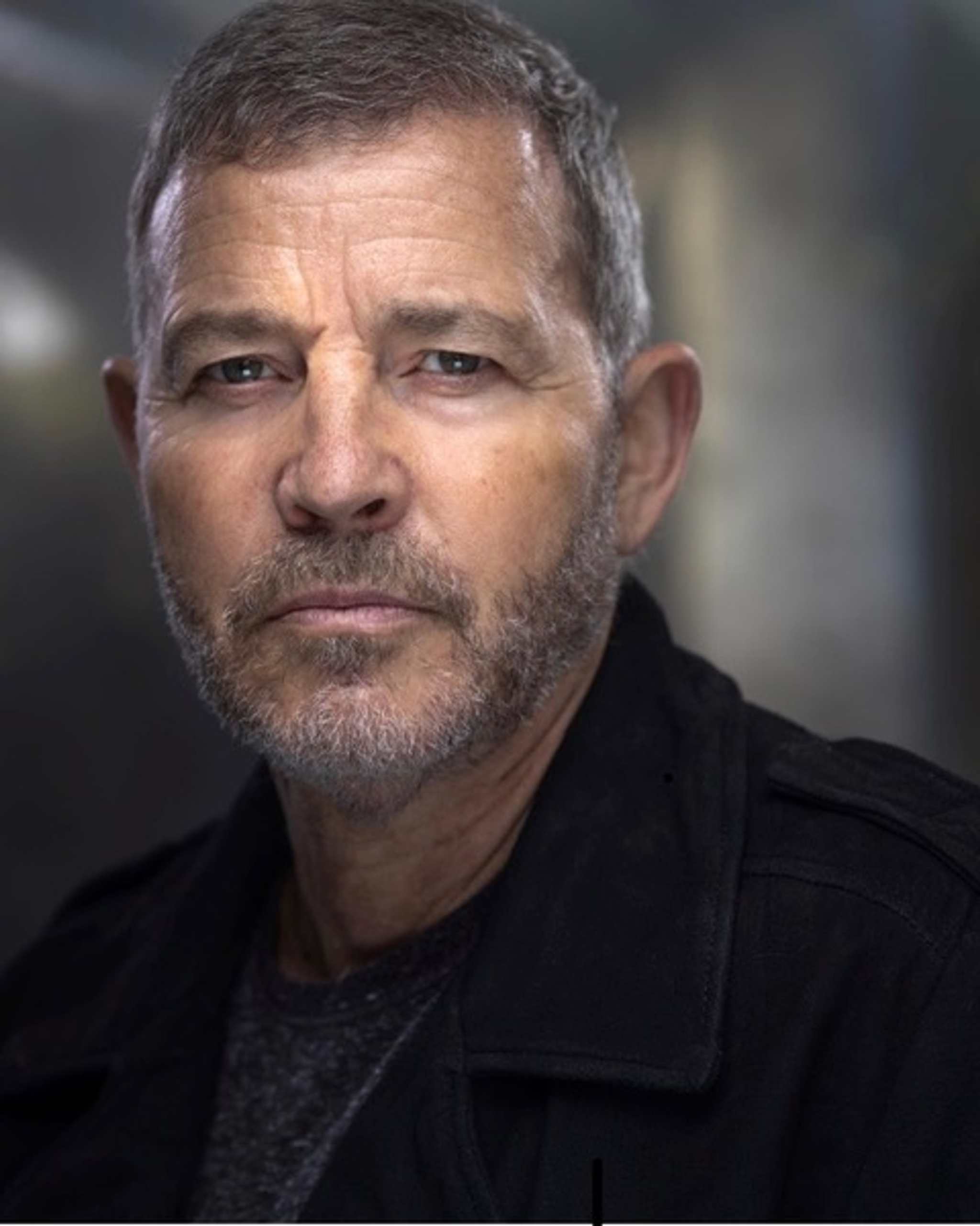 A portrait of actor Andrew Lewis seen from the shoulders up. He has a short beard and wears a dark blazer. A light highlights his forehead and cheek bones. 