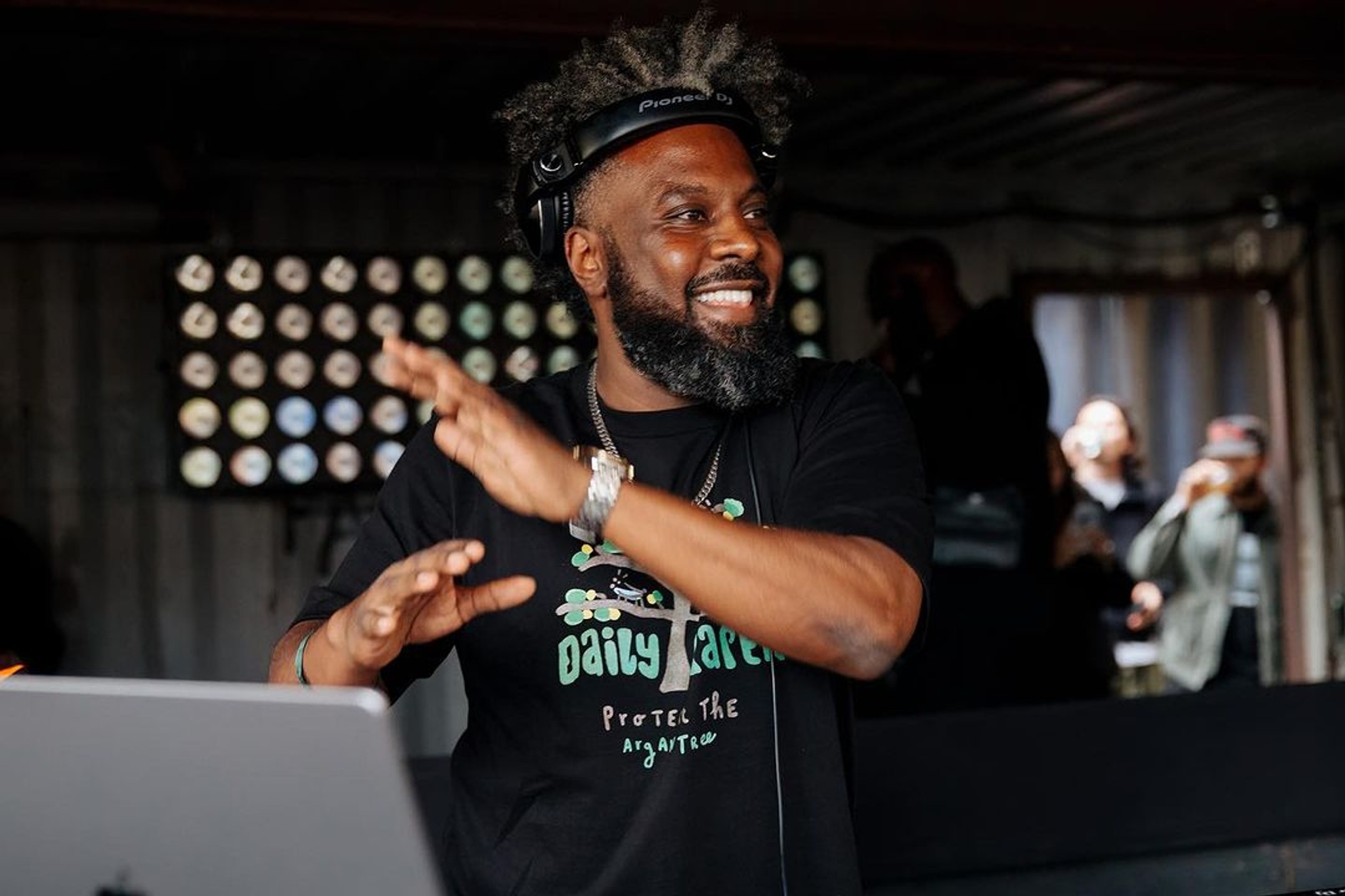 A Black man stands with arms raised as if mid-dance. He is smiling looking to the side and stands in front of an open laptop computer