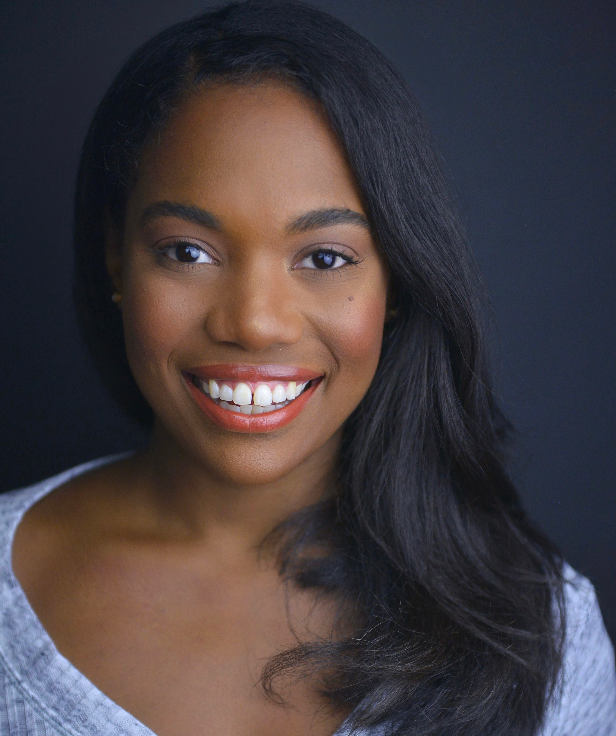 A Black woman smiling at the camera picture from the chest up. Her hair is brushed around one side of her head to swoop over her left shoulder.