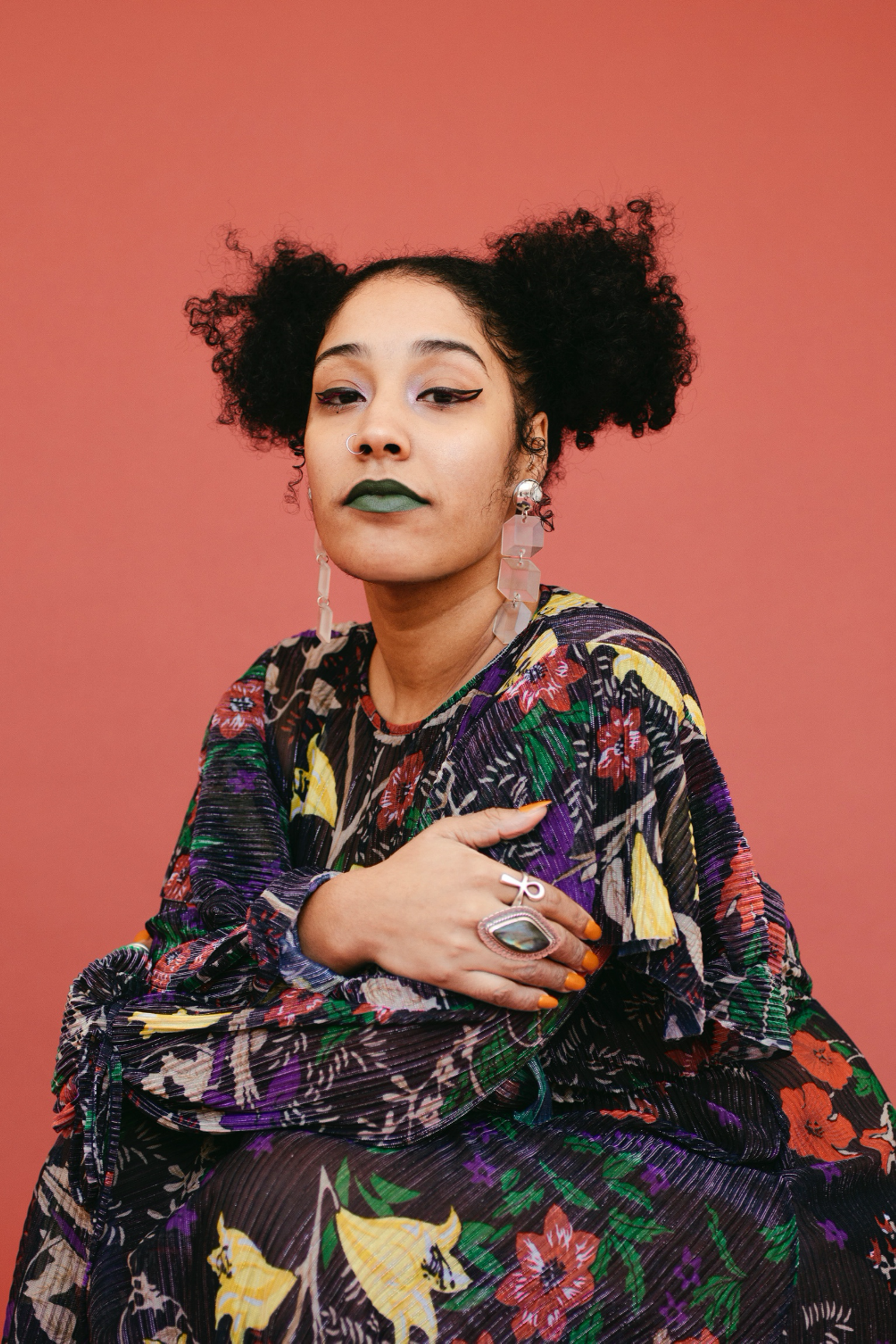 A photo of Suhaly Bautista-Carolina wearing a floral print dress, large rings, green lipstick, and long earrings. 