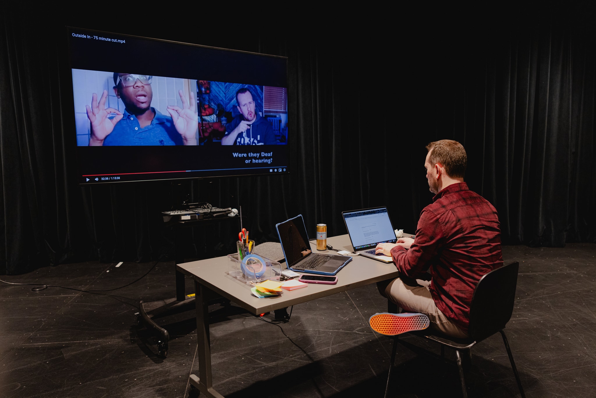 The artist Garrett Zuercher is seen from behind seated at a wide table in front of a large tv monitor. The desk is covered with a laptop and a stack of post it notes. Garrett is training his attention on the tv screen which shows a split screen of him and a Black Deaf artist in a recording of a virtual meeting.