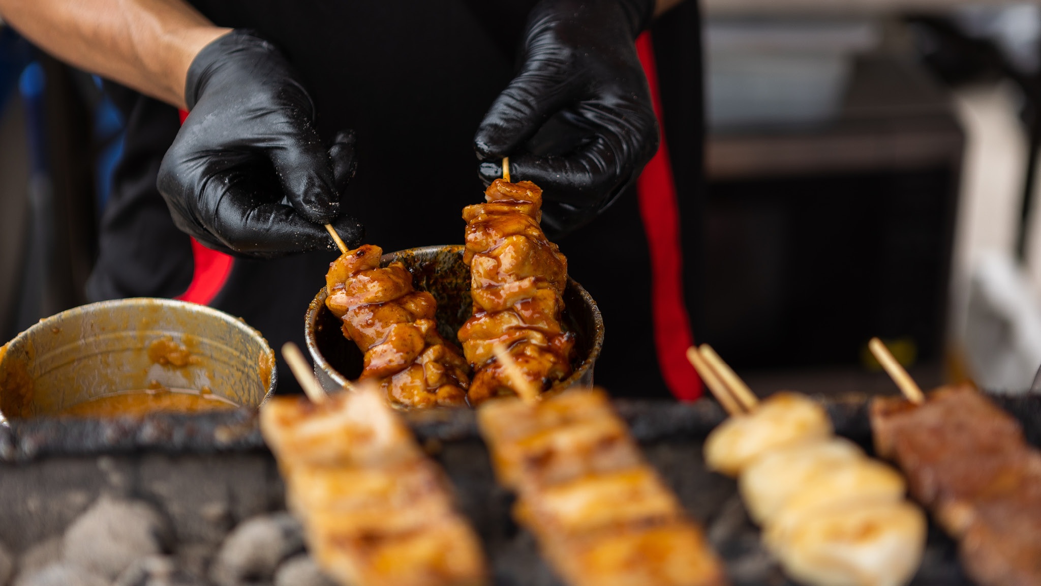 A close-up of a person's hands in black latex gloves holding two meat skewers above a grill. In the foreground, a row of skewers sit on the grid. 