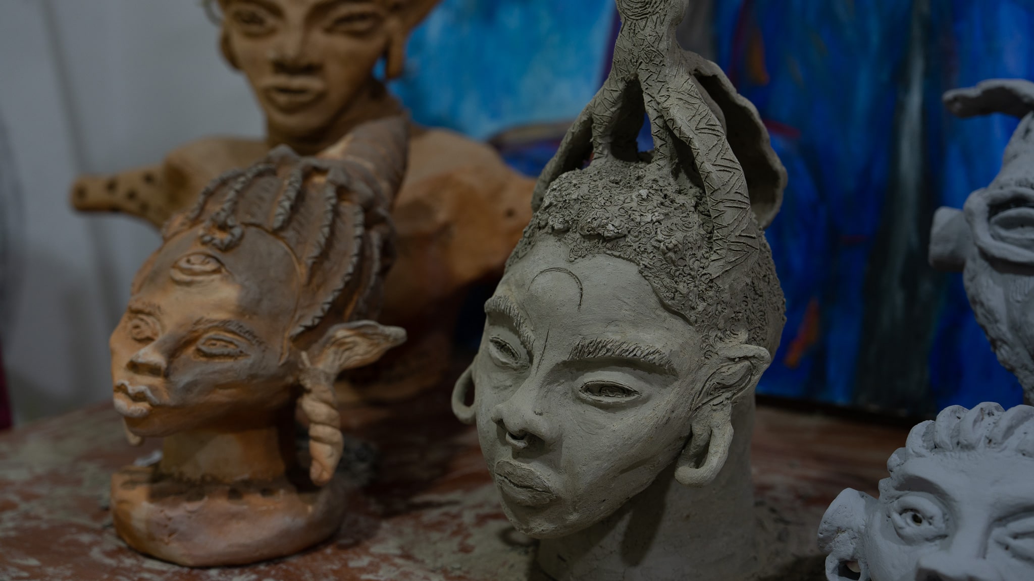 Ceramic bust sculptures are seen lined up on a table in artist Chelsea Odufu's studio. The busts are created using African motifs and depict women's heads. They have braided hairstyles and the central sculpture in the image has a moon shape on her forehead and wears a nose ring in her septum. The sculptures are in clay tones of gray and terra cotta. 