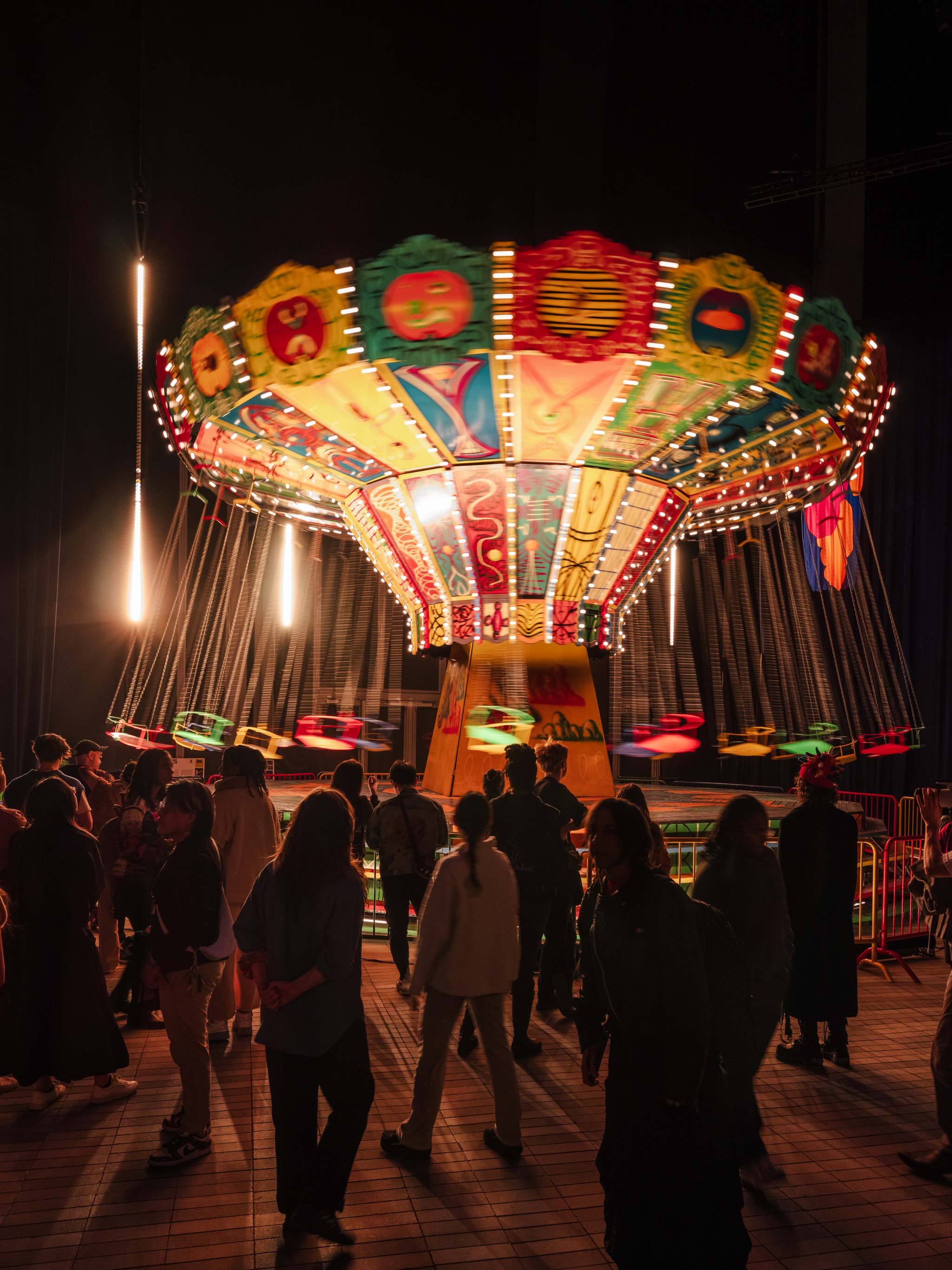 A crowd of gallery visitors look on in awe as a painted chair swing ride by Kenny Scharf is illuminated and spins around. The panels of the ride's interior are painted in bright colors, and the brightly colored chairs swing jubilantly on their chains.