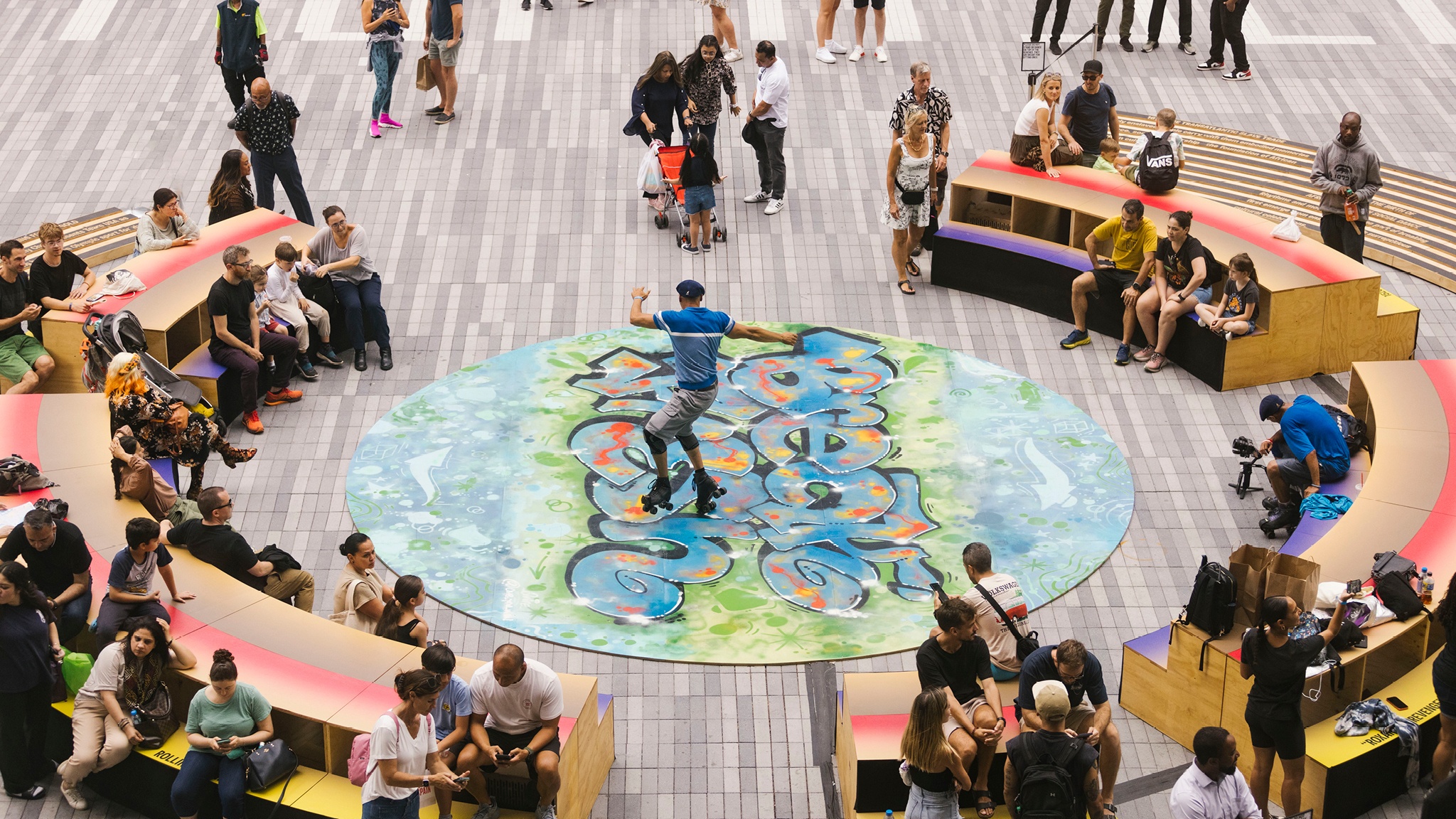 A crowd gathers on an outdoor plaza around a dancer performing on a wide, round painted surface. The surface has been painted in a graffiti style in a neon green and blues.