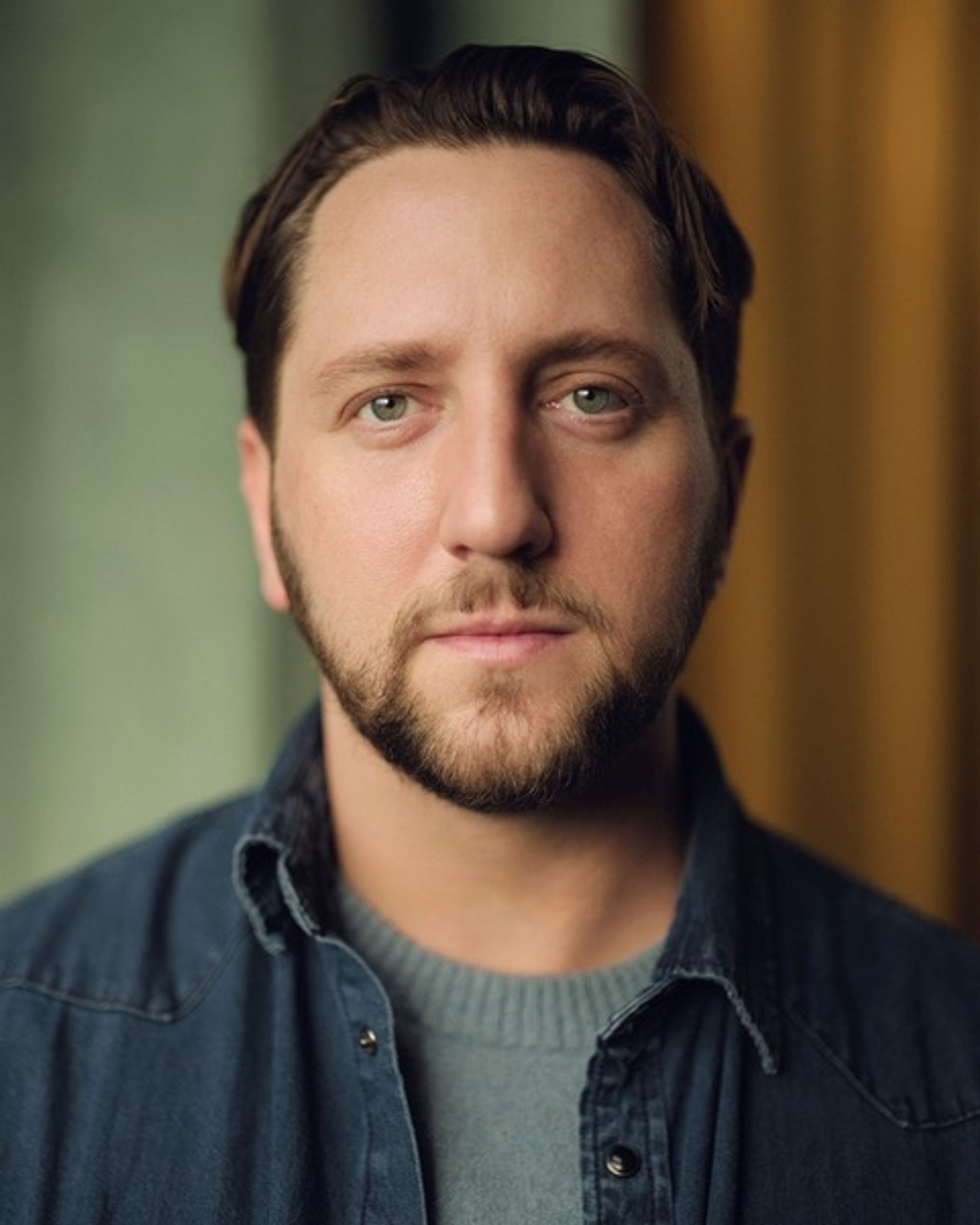 A portrait of actor Adam Silver, a white man with brown hair parted to the side and a beard and mustache framing his chin. He looks to the camera without smiling. 