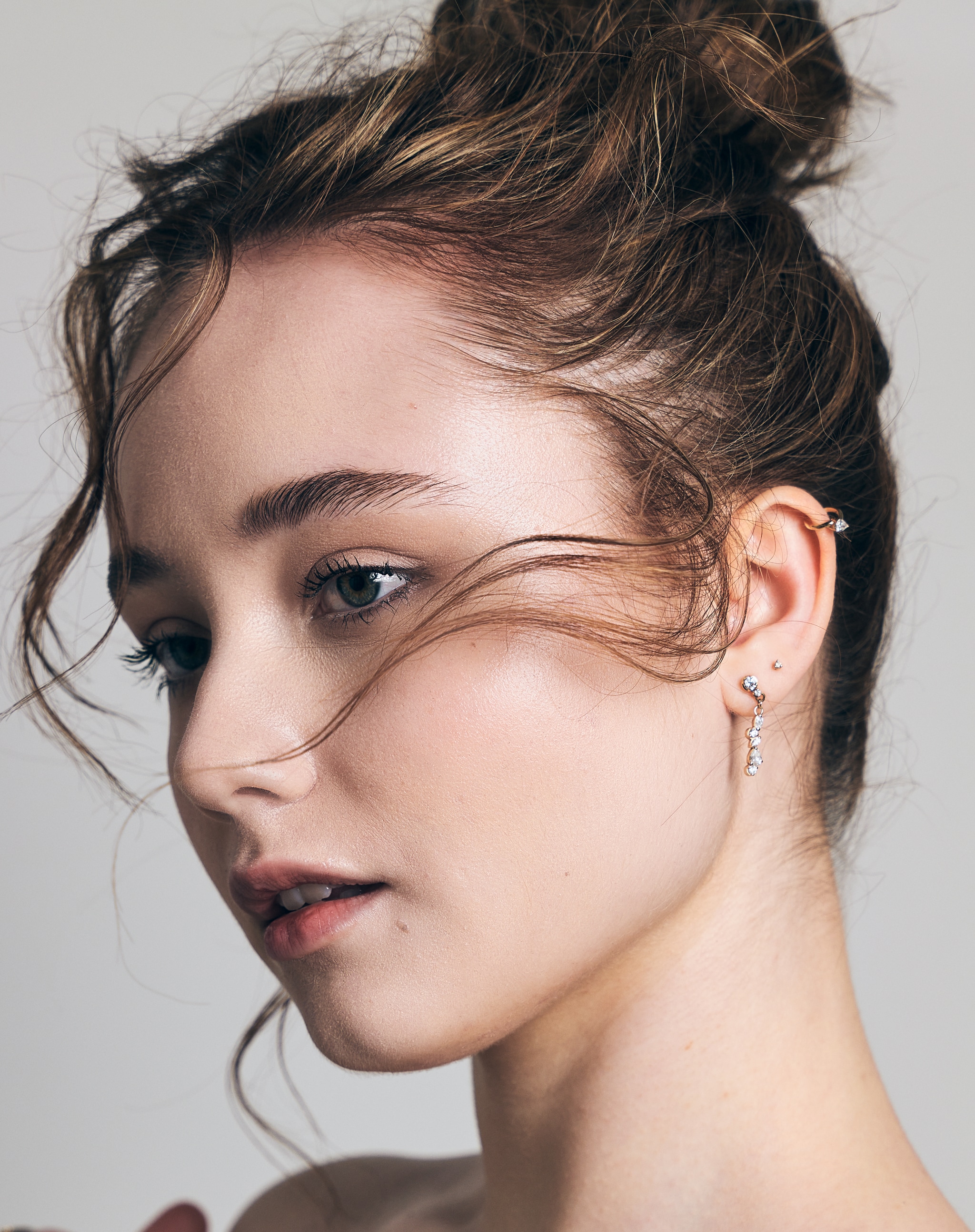 A photo portrait of actor Ruby Stokes. Ruby's head is seen in three quarters profile from the left. Her light brown hair is pulled up in a loose bun with wisps across her cheek. She looks down away from us with a contemplative look on her face. 