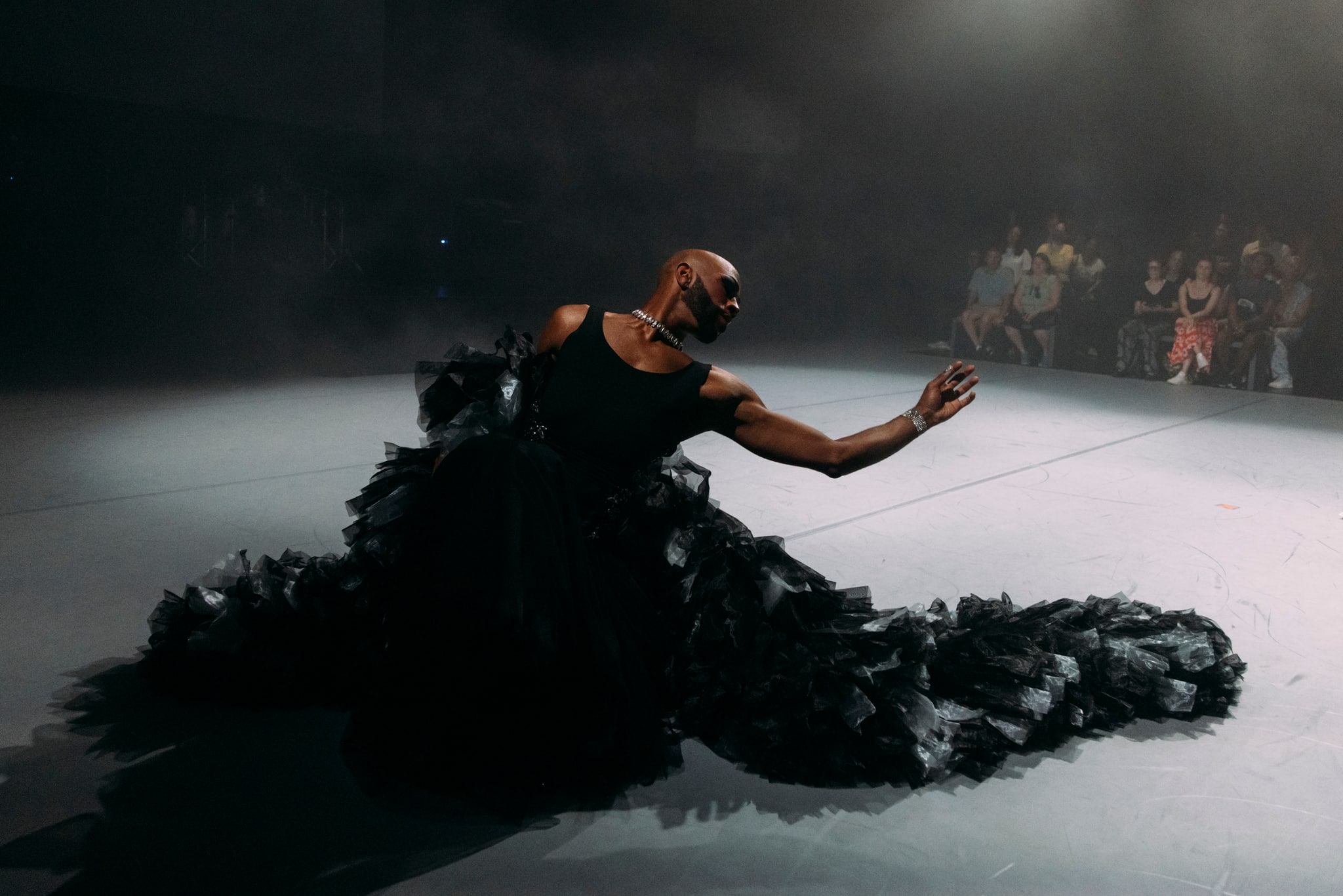 Cain Coleman, a Black performer and artist, dances on stage during the performance of "New Information." Cain wears a ruffly, billowing black gown, and seems to sink into it on the floor, one arm gracefully outstretched.
