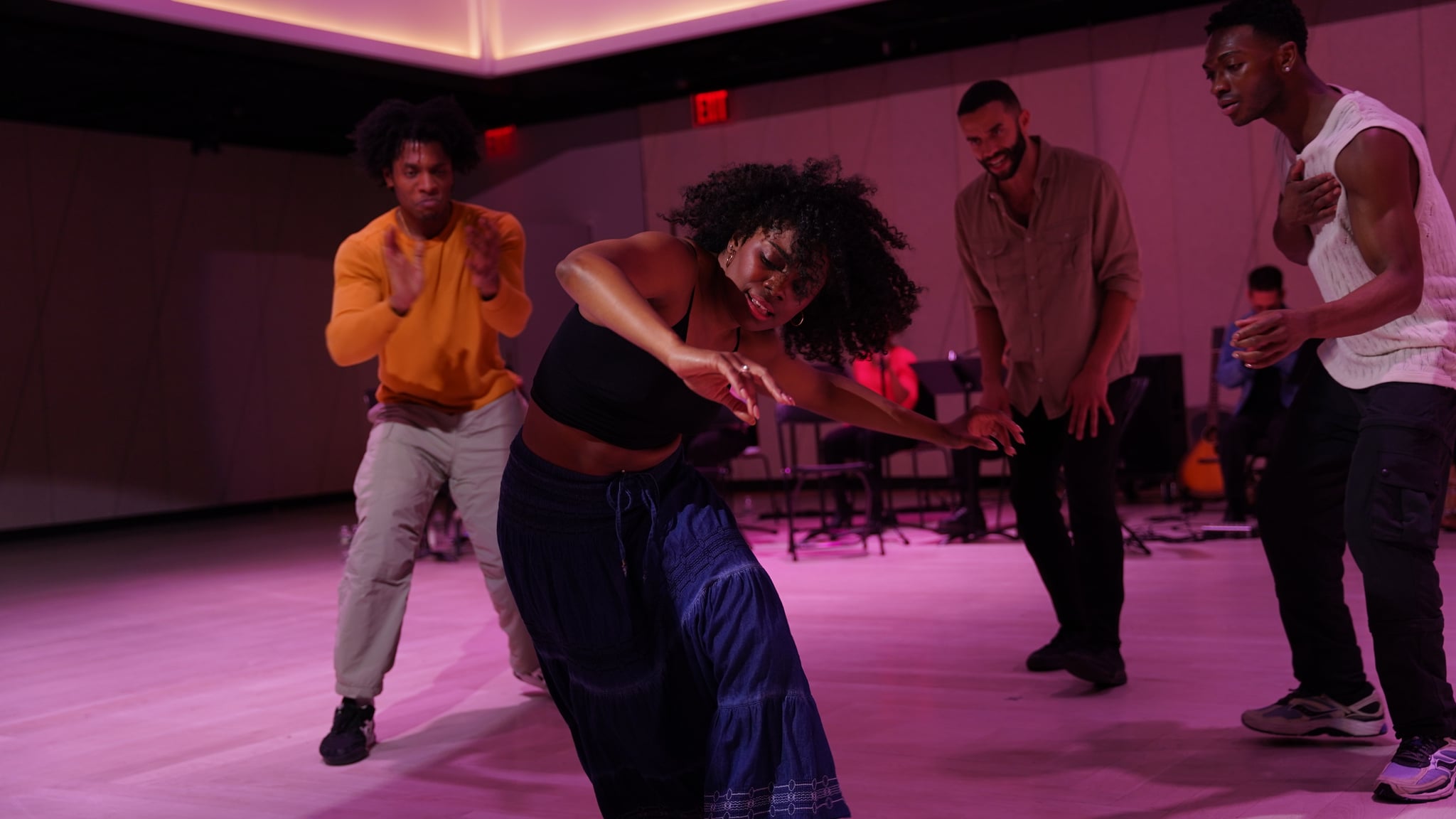 A Black woman dancer curves her body dramatically to the left with one arm oustretched, while three Black men dancers stand in the background clapping and watching in encouragement. The dancer in the foreground wears a black tank top and a long dark blue skirt. 