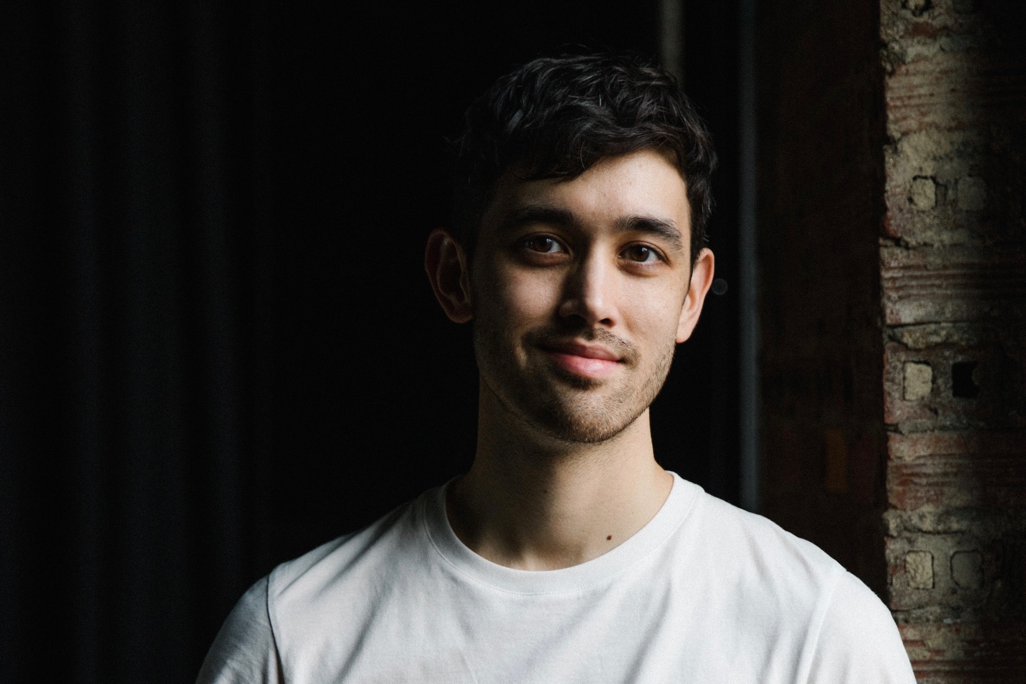 A photo of the artist Benjamin Akio Kimitch. Kimitch wears a white shirt and light falls on the left side of his face. The left background of the photo is in darkness. 