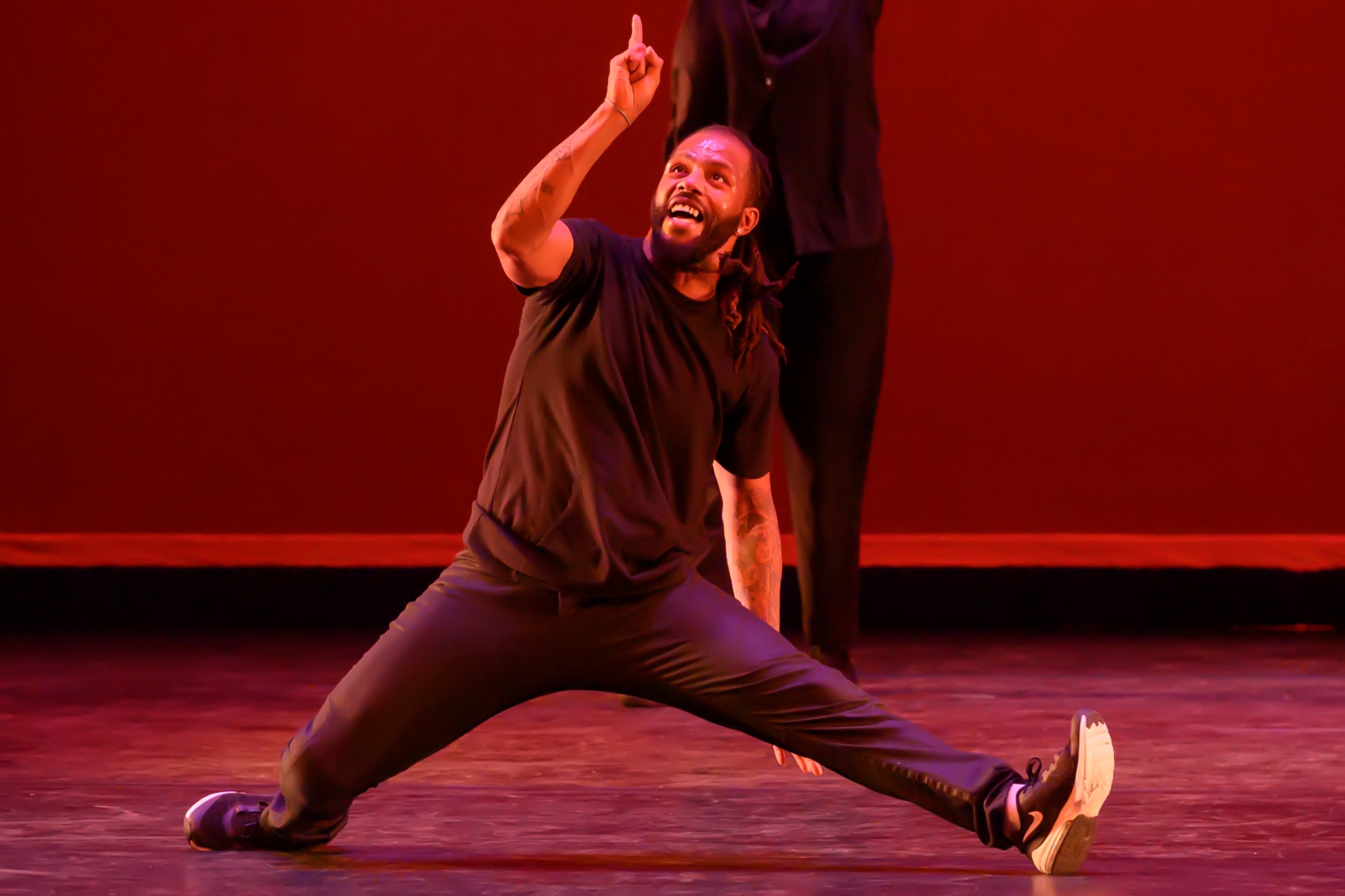A Black dancer points upward while mid-split on a stage. 