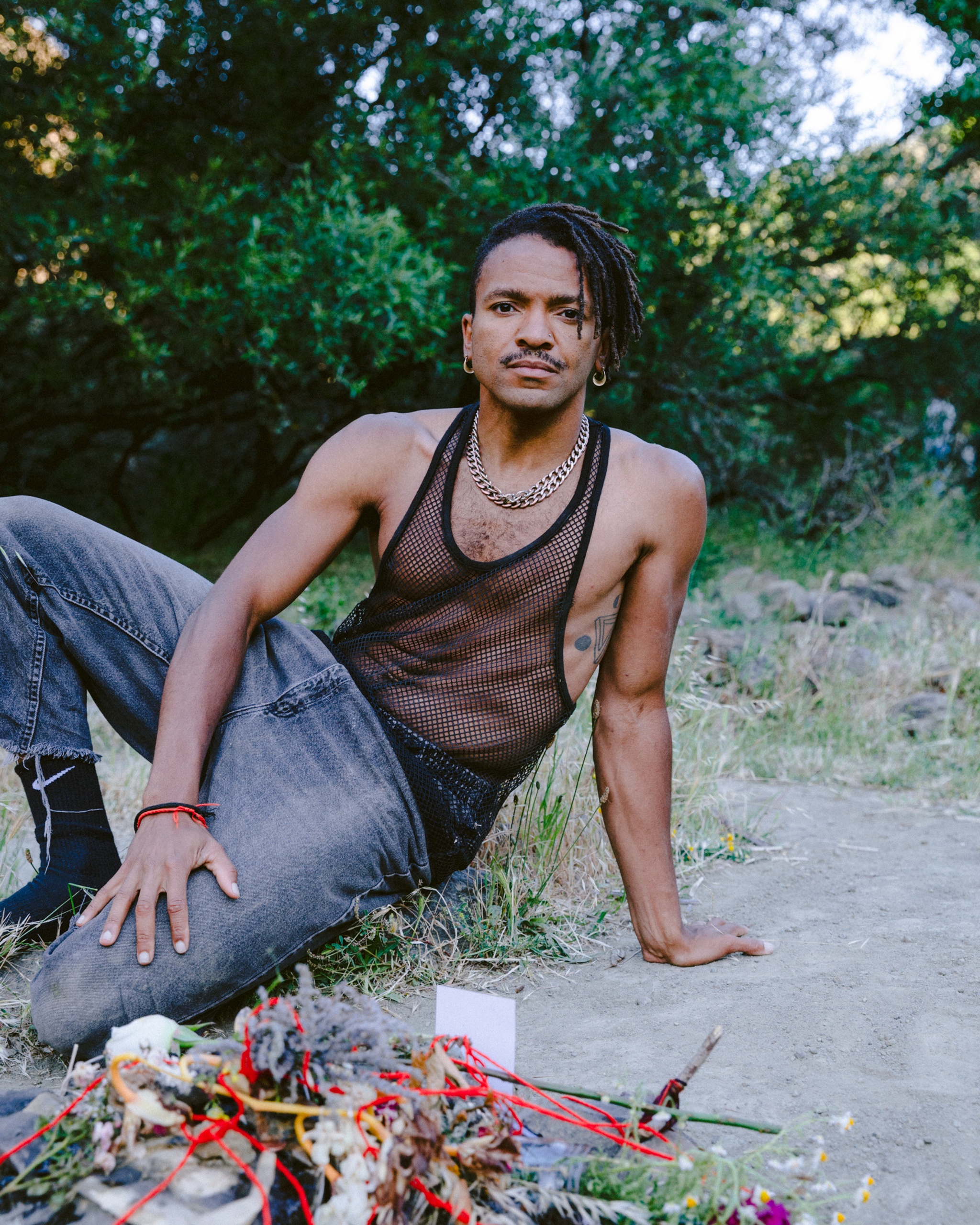 The artist Gabriele Christian, a Black nonbinary person, reclines outside on the ground. They lean back on one arm with the other draped over their leg and lap. They wear a mesh black tank top and jeans. In front of them is a tangle of brightly colored string, flowers, and other materials. Behind them is a screen of leafy green bushes and trees. 