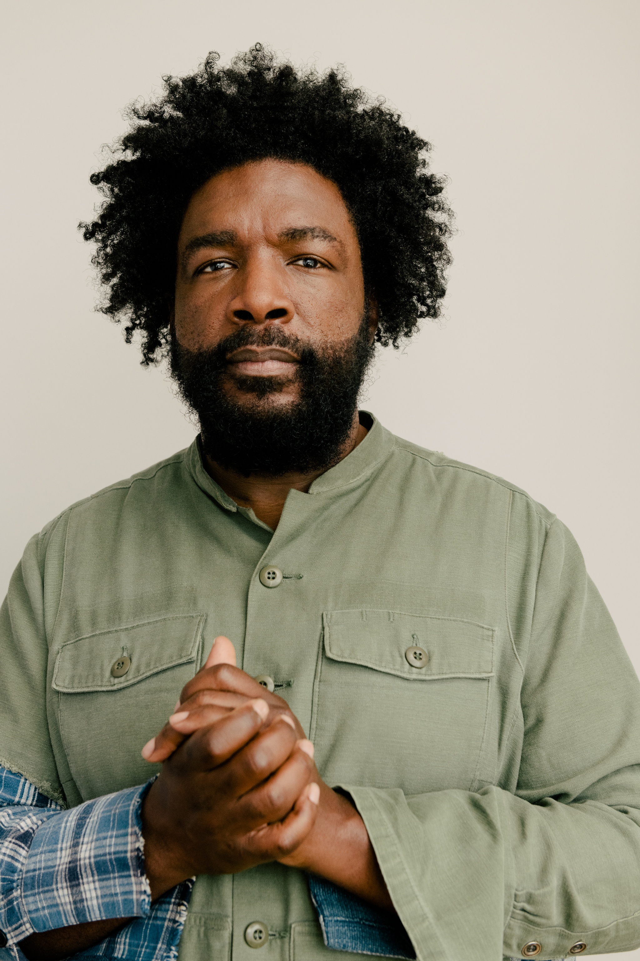 A portrait of the musician Questlove, a black man with a beard who is clasping his hands in front of his chest. He wears a pale green shirt with blue plaid cuffs. 