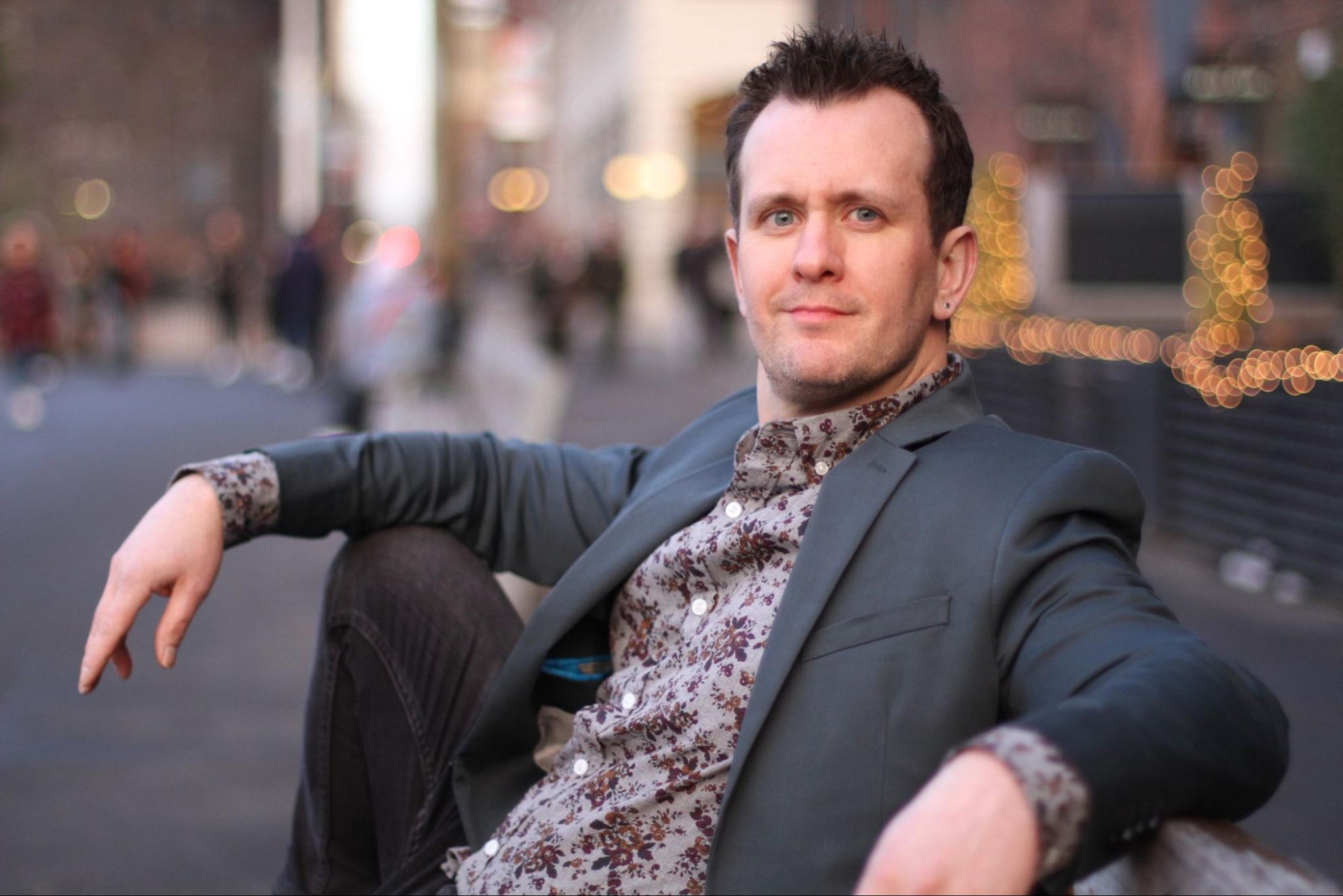 A portrait of Garrett Zuercher, who sits on a bench along a city street. He has one arm propped on his knee and the other draped over the back of the bench. He wears a blazer over a floral print shirt and turn to look toward us. Photo by Jo Welch