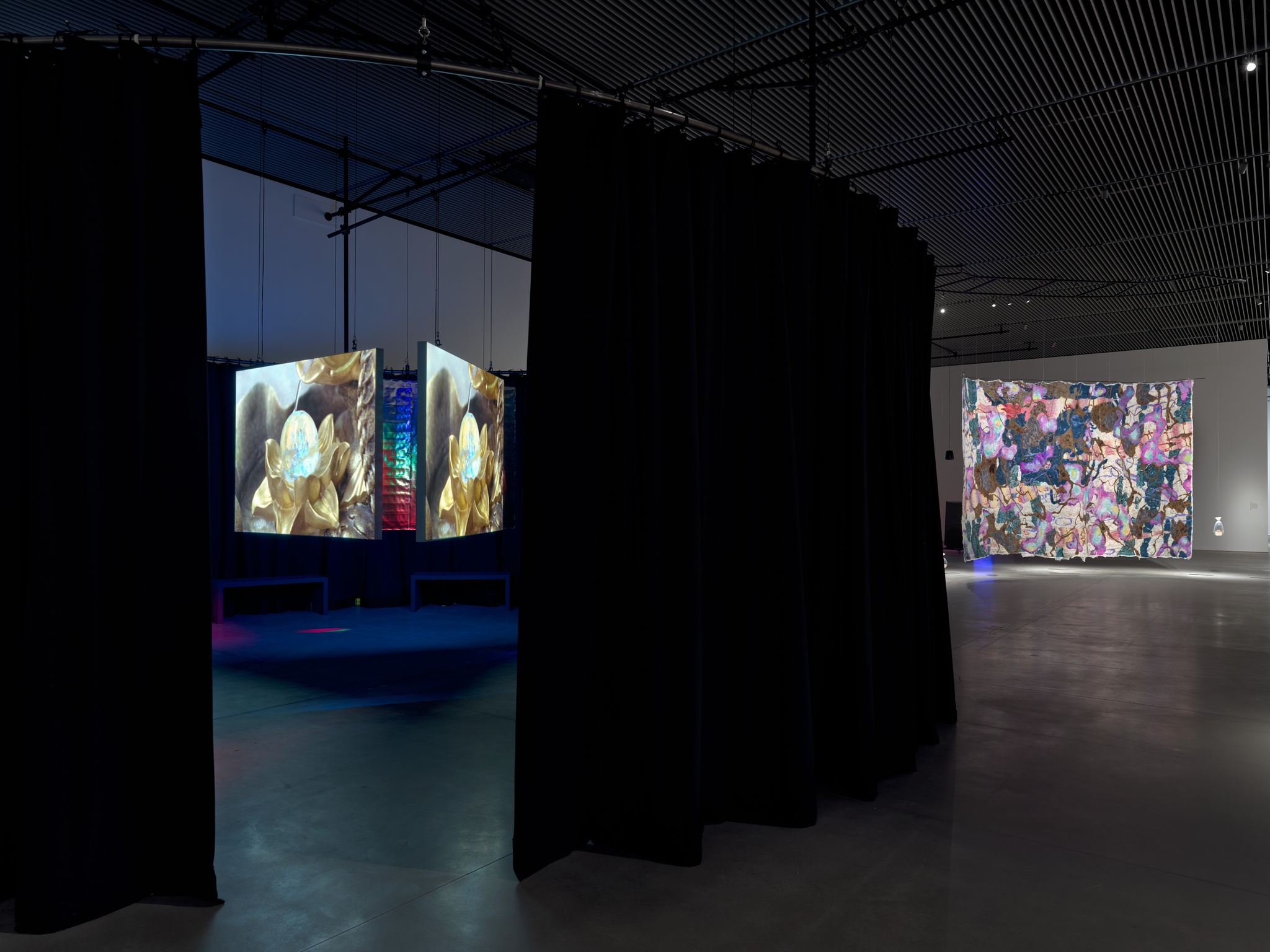 An art gallery with a set of three video screens inside a curtained area on the right and on the left in the background a hanging multicolored felt tapestry