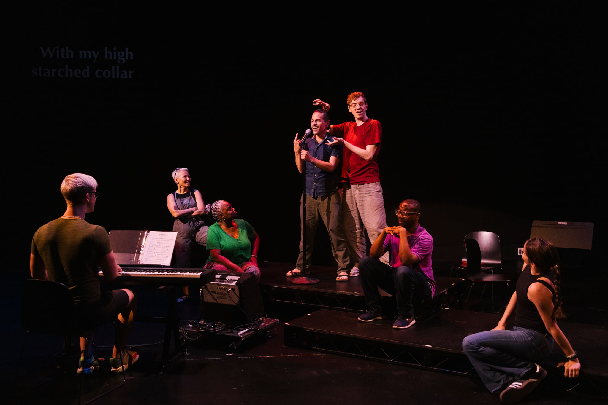Six Deaf performers appear on stage in Garrett Zuercher's "Inside/Look." One central performer stands at a microphone while the others look toward him. In the foreground, one of the performers is seated playing a piano with their back to us.