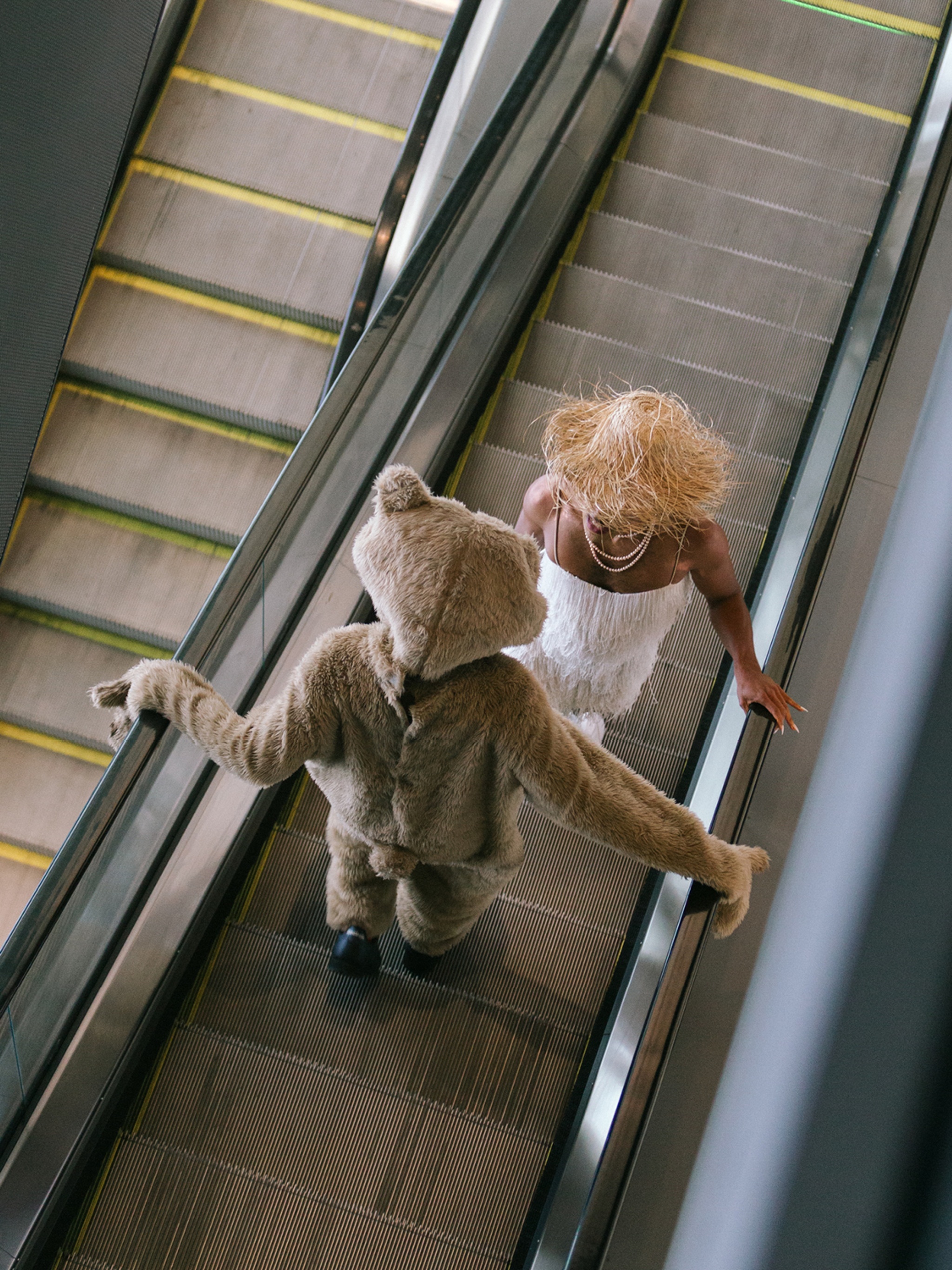 The performers NIC Kay and Gabriele Christian are seen from above riding an escalator. NIC is in a full-body brown bear costume, and Gabriele's face is hidden by a wide-brimmed straw hat. 