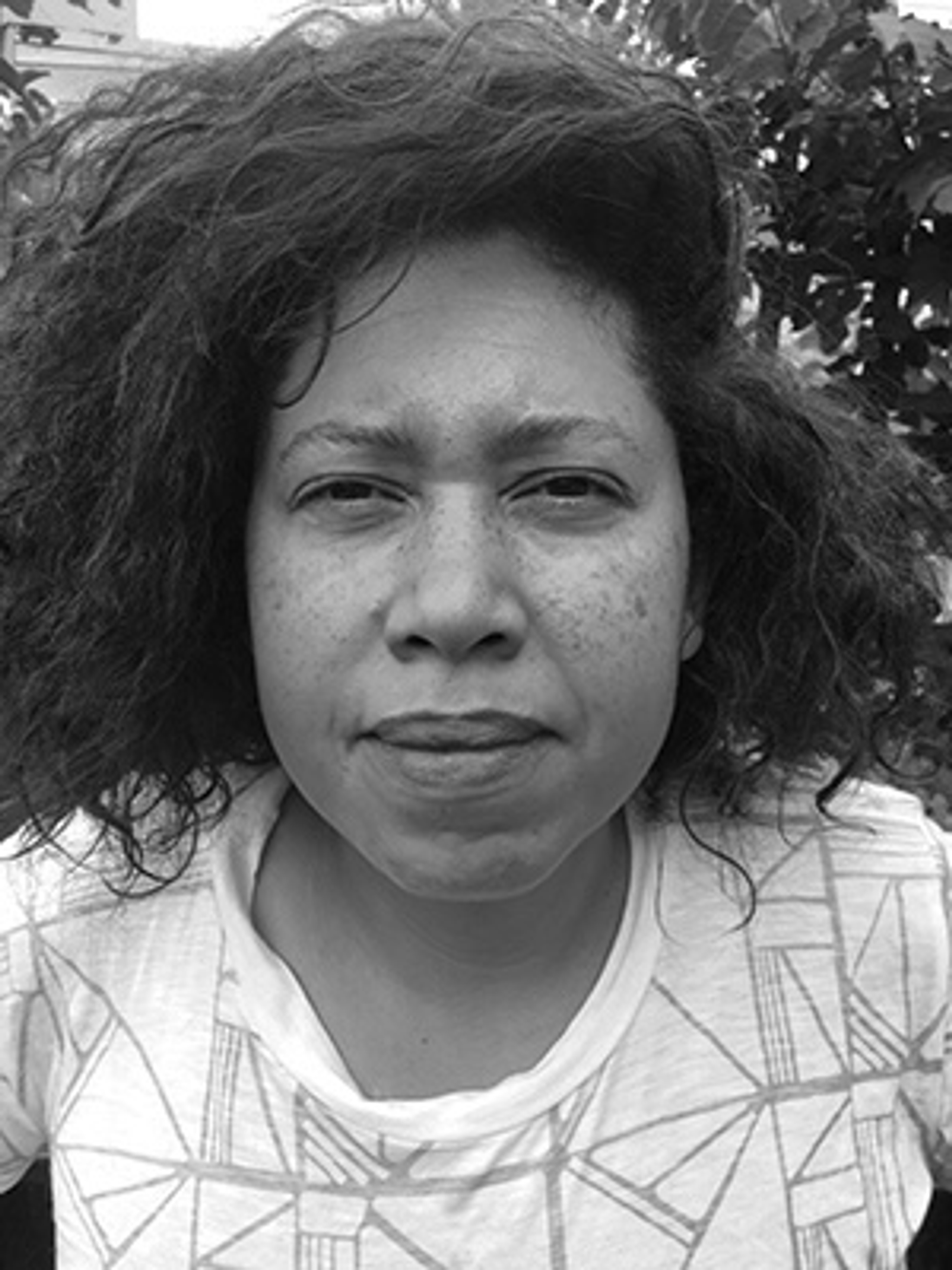 A black-and-white photo of Abbey Williams with her hair pushed over the right side of her head. Williams wears a white t-shirt with a geometric pattern print. 