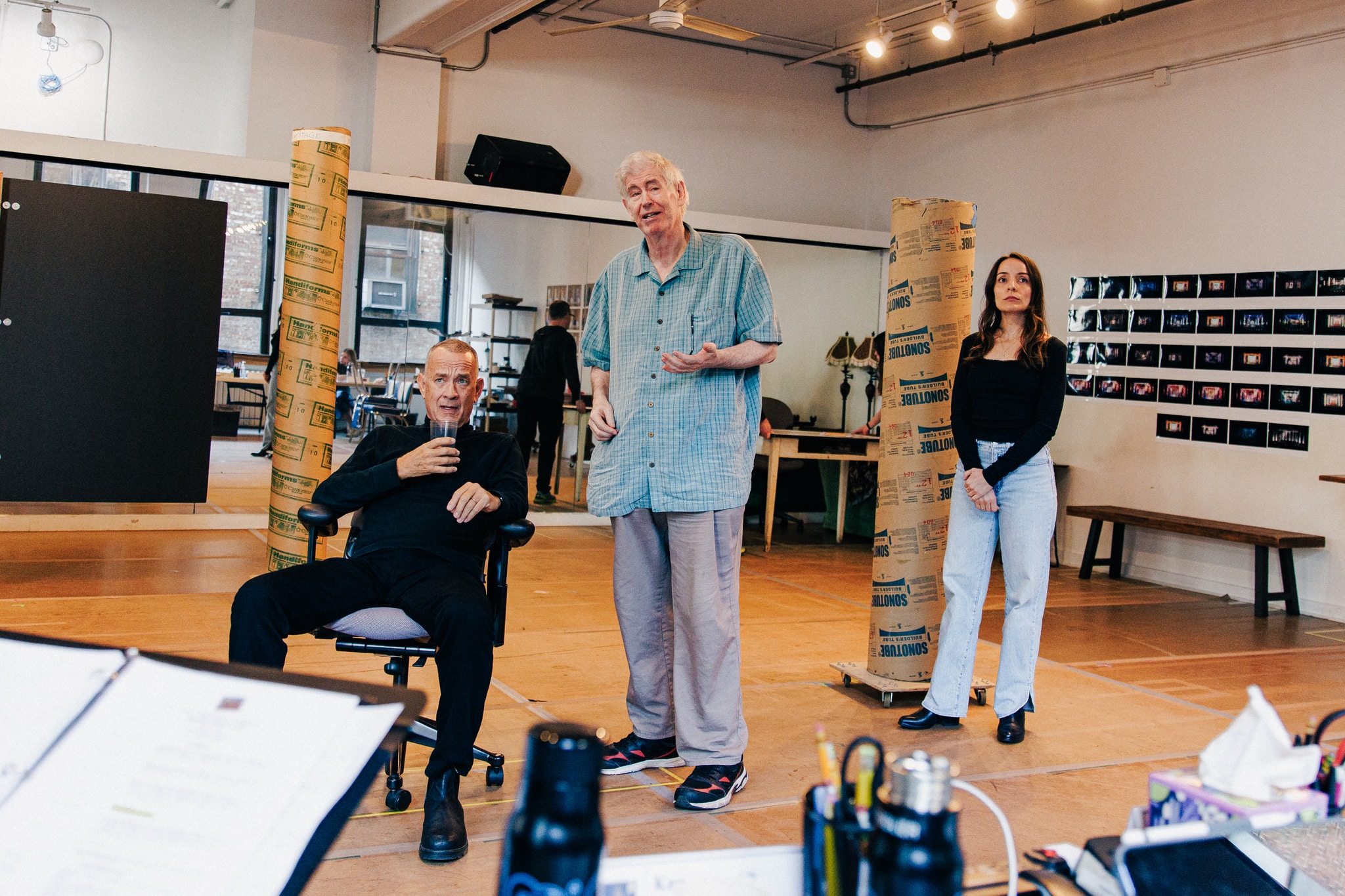 Actors Tom Hanks, Paul Murphy, and Jamie Ann Romero rehearse in a brightly lit dance studio. Tom is seated with Paul standing at his side. Jamie Ann stands slightly in the background to Paul's left looking off in the distance with hands clasped in front of her. 