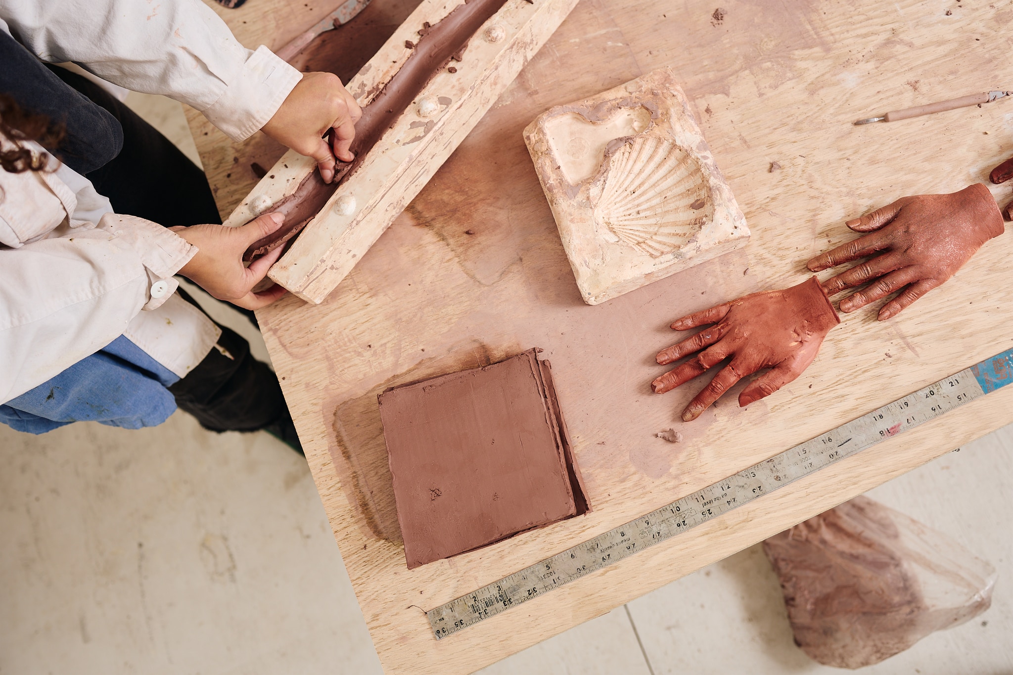Artist Patricia Encarnación's hands are seen from above in the top left corner of the photo as she touches sculptures in progress on a table in her studio. The table includes the mold for a seashell sculpture in the shape of a large clam shell and two sculptures of human hands in an orange-brown earthenware color. 