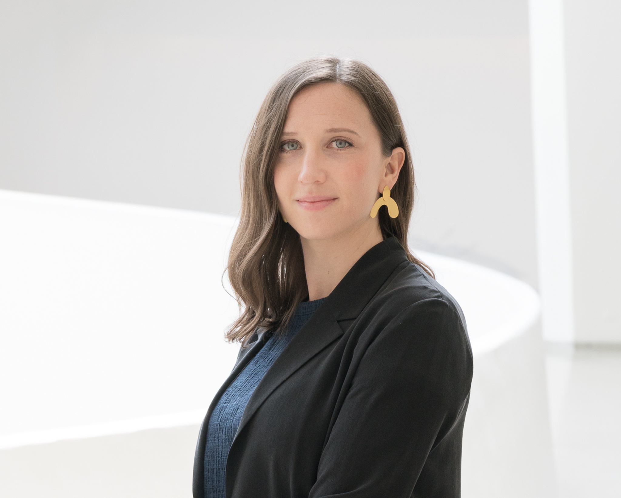 A photo of Susan Thompson in a black blazer and wearing gold earrings, posing against the white background of the Guggenheim Museum's rotunda. 