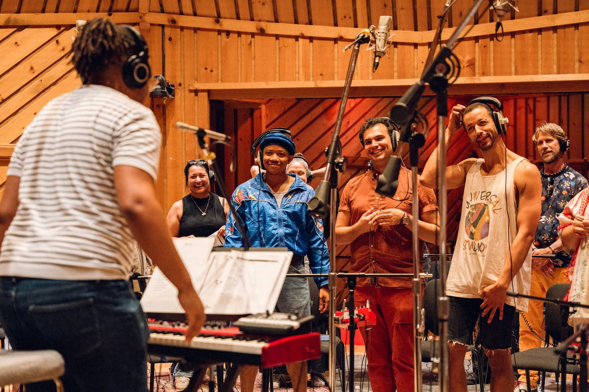 A row of choir members stands in front of a row of microphones, facing Troy Anthony (who we see from behind). The choir members where t-shirts and tank tops, casual summer attire, and are smiling and laughing as they pay close attention to Troy.  