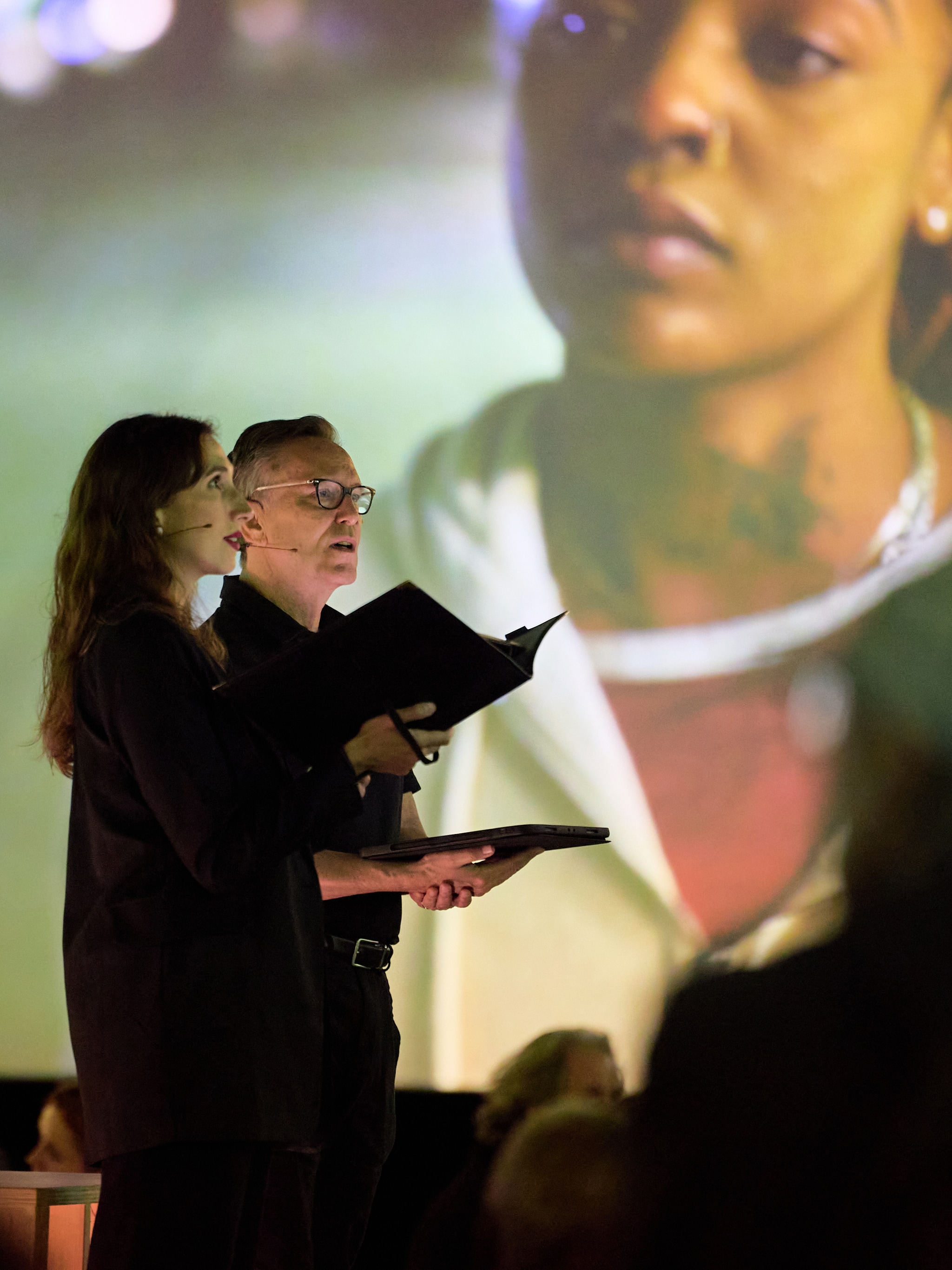 Two chorus members dressed in black sing while silhouetted against a film screen with the projected image of a Black woman behind them. The woman on screen wears a gray jacket over a red shirt and looks down to her right. 