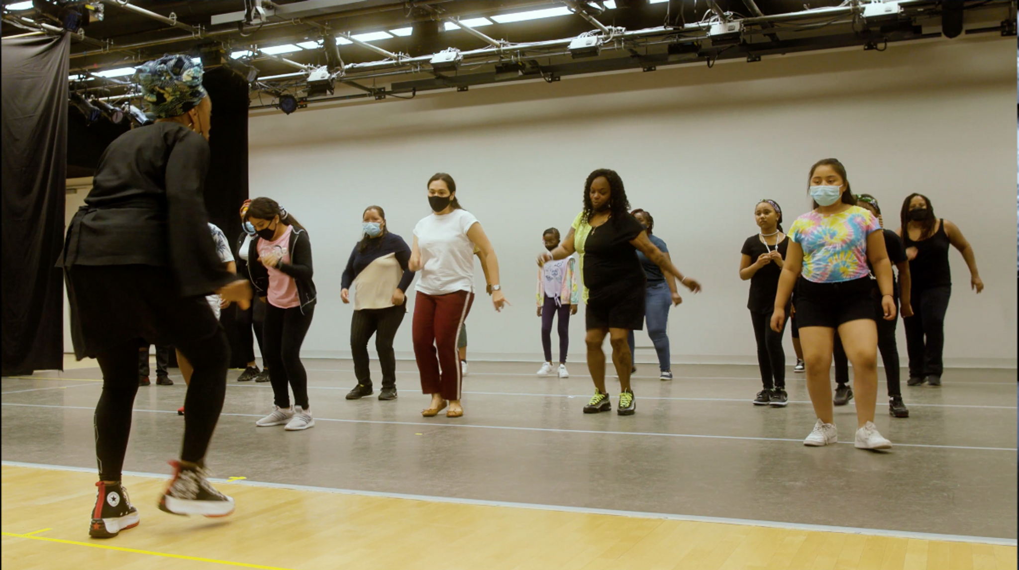 A group of people in casual athletic wear dancing together in a dance studio, led by an instructor dancing in front of them.