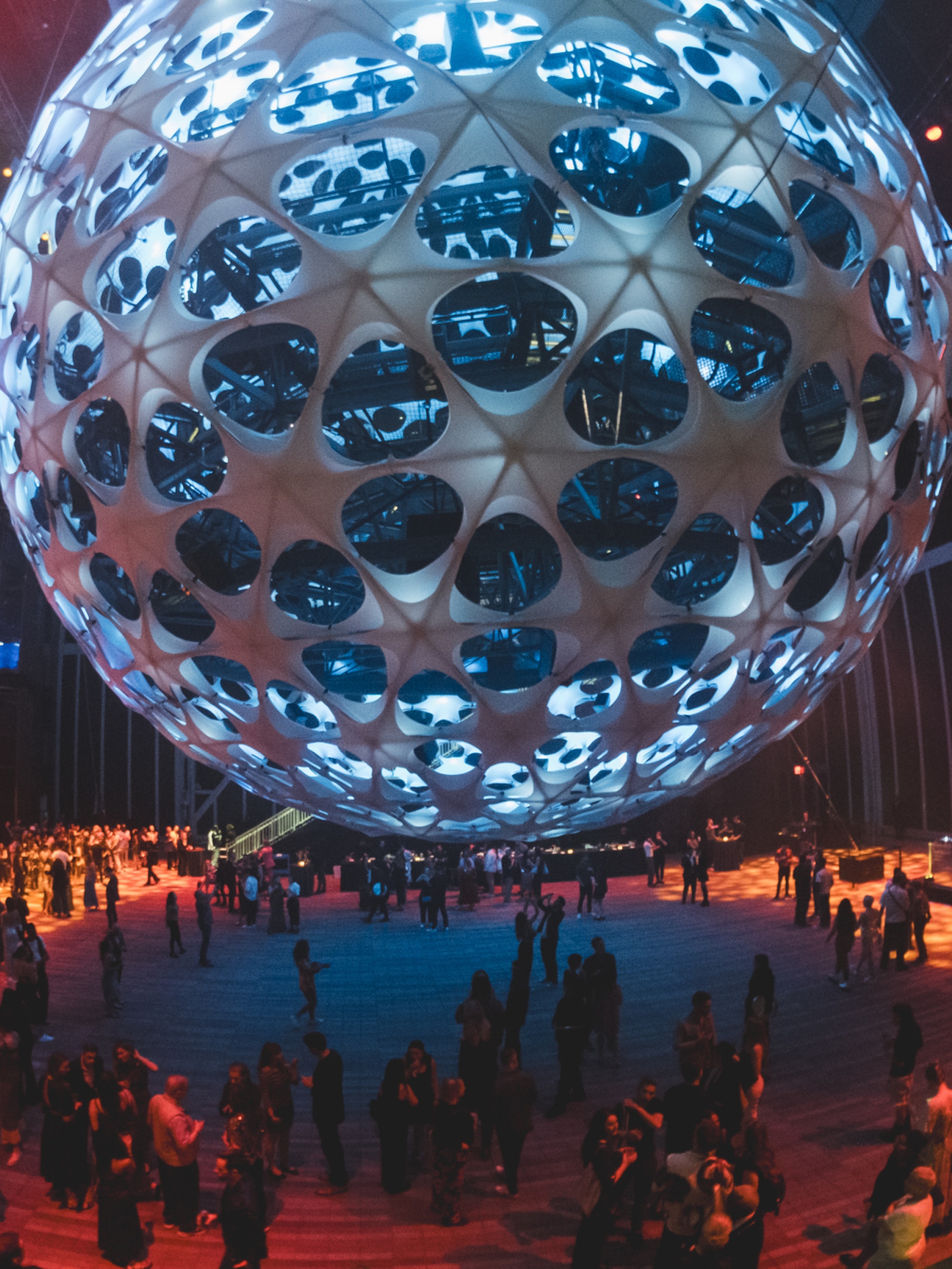 A crowd gathers beneath a large spherical concert hall suspended in air in a vast performance space. The sphere can be seen into through large apertures that make it resemble a wiffle ball. It is lit in pale blue light, and golden light illuminates the ground around its edges. 