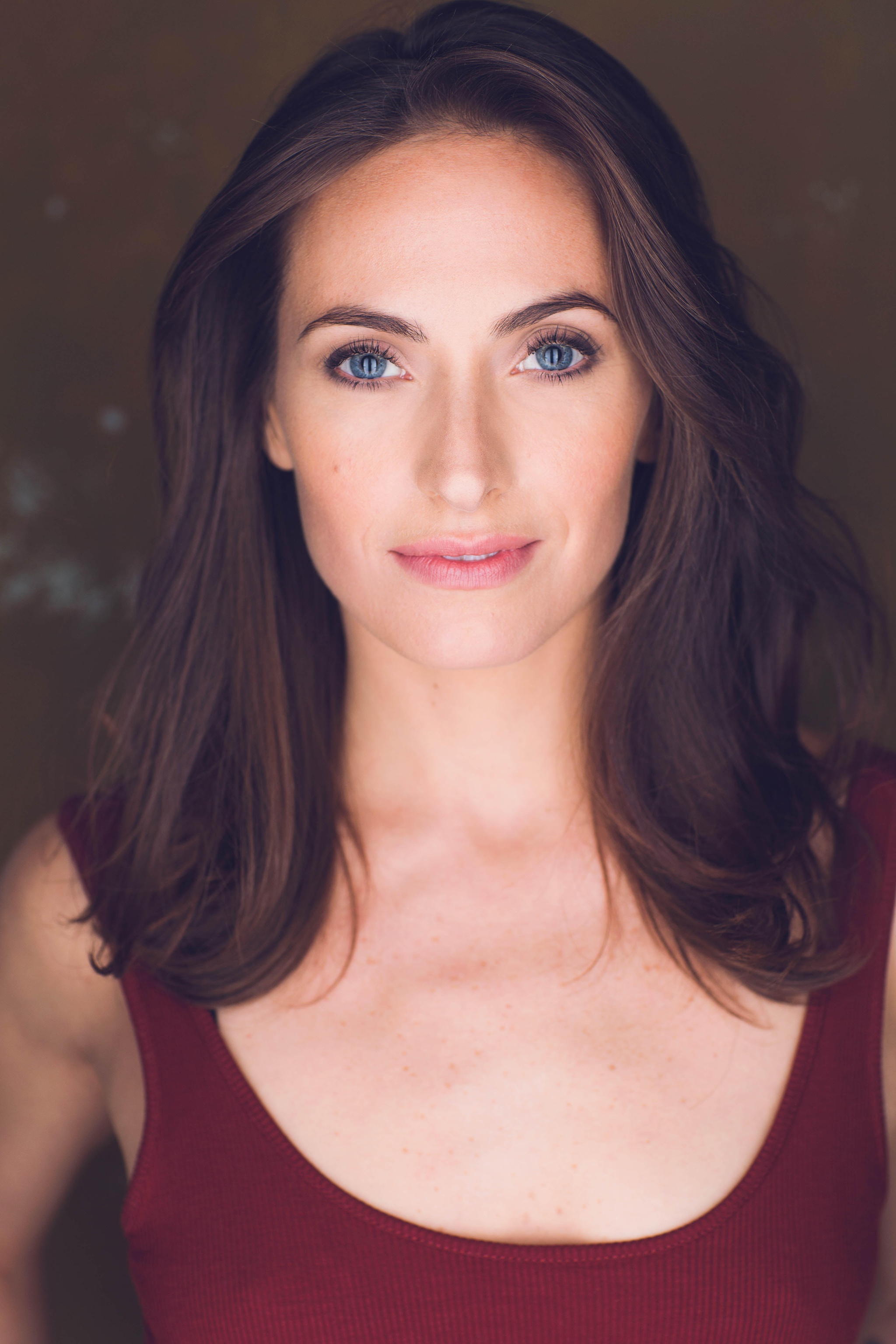 A portrait of Stephanie Sutherland against a dark brown neutral background. Stephanie has long brown hair that frames her head to her shoulders. She wears a tank top and looks directly at us, smiling without opening her mouth. 