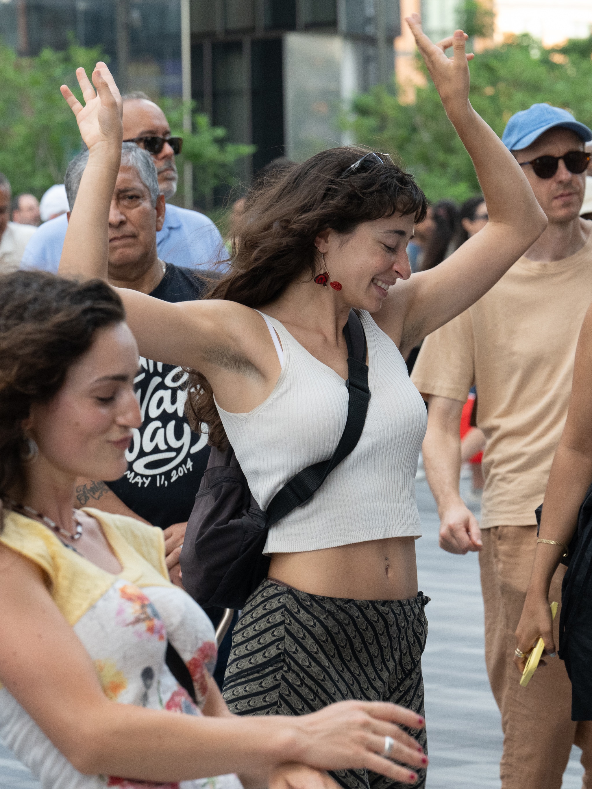 A young white woman is seen dancing with arms raised above her head. She is outside on a sunny summer day. She wears a white tank top and has brown hair that falls down her back.