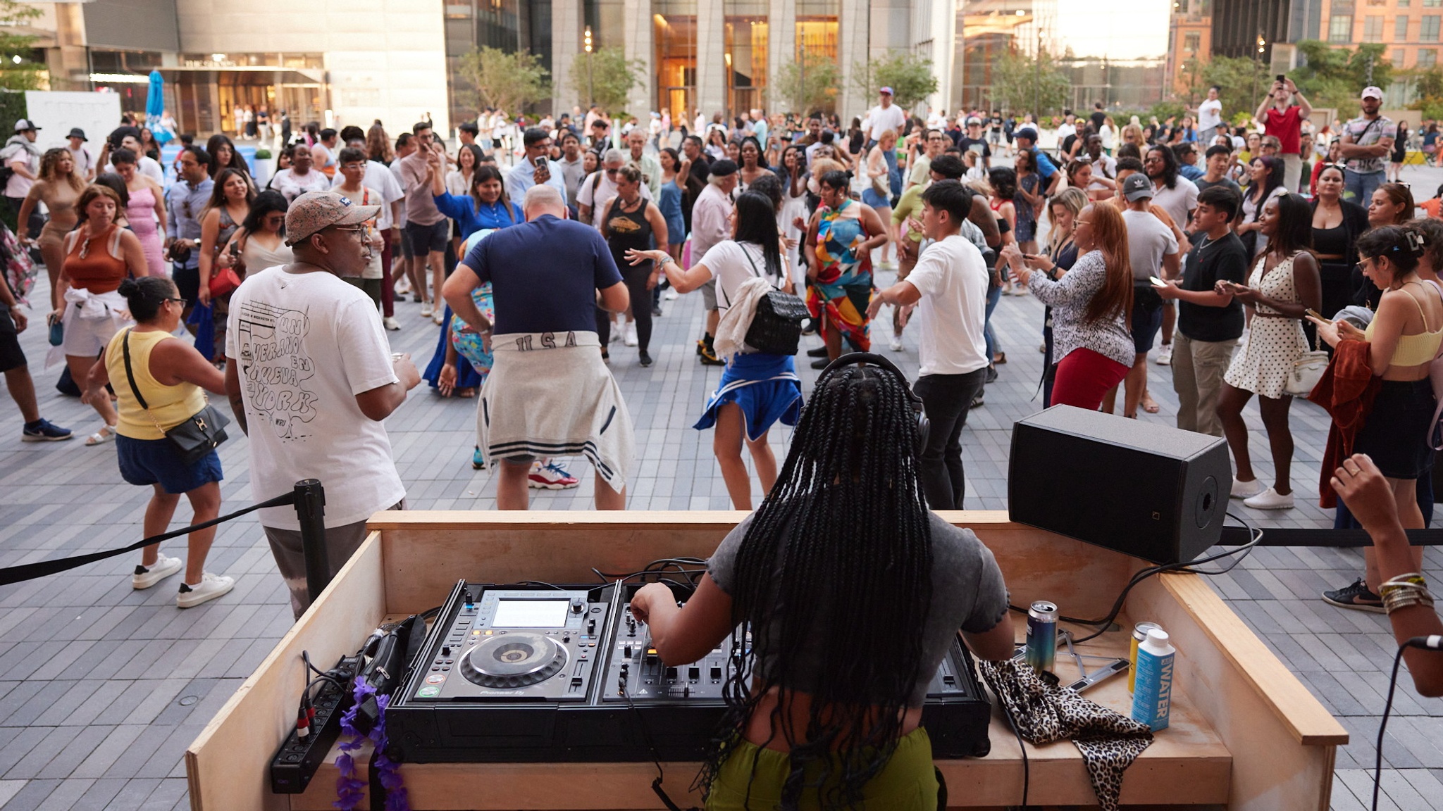 A DJ is seen from behind in a DJ booth outside on an open Plaza. In front of her a crowd dances on a warm, sunny, summer day. 