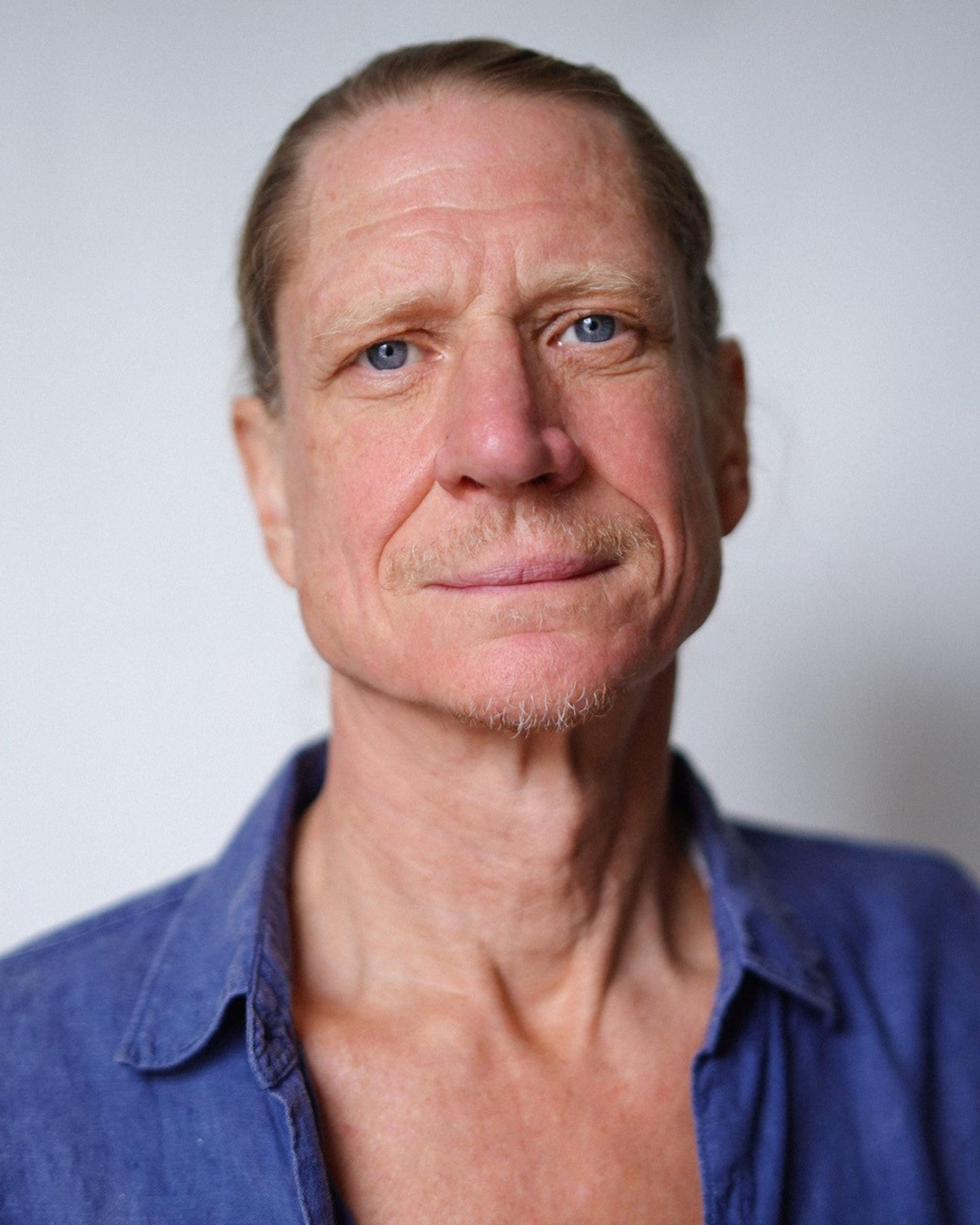 A photo portrait of actor Jerry Killick. Jerry faces us directly and is pictured in a tight shot from the chest up. He has light brown hair pulled back behind his head and smiles warmly with mouth closed and a sympathetic look in his eyes. He wears a blue button down shirt. 