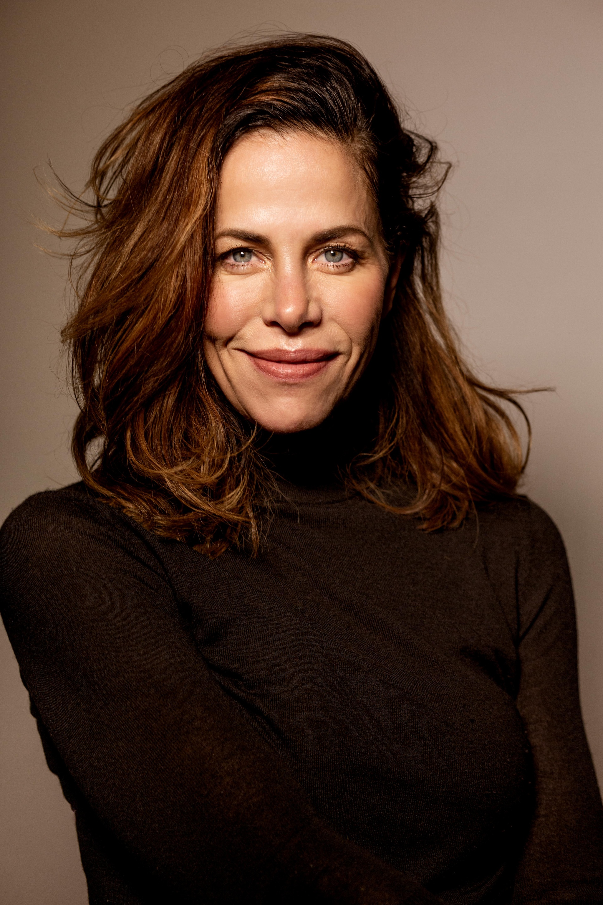 A portrait of actor Jennifer Regan. She is a white woman with wavy brown hair to her shoulders and wears a black turtleneck. 