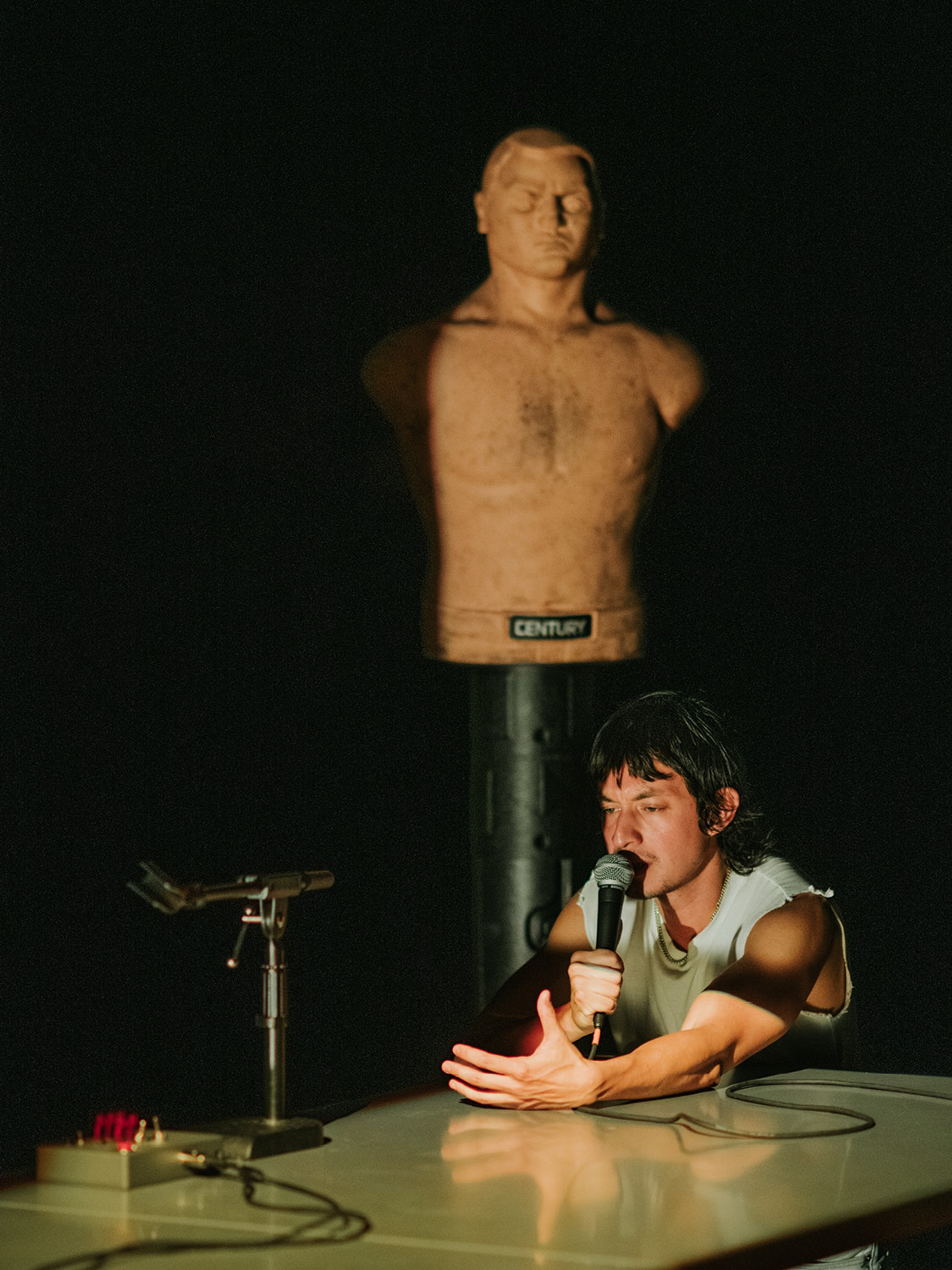 Kyle Dacuyan performs on stage, seated at a table with a microphone held to his mouth. He reaches one arm out across the table in front of him. Behind him looms a sparring dummy. 