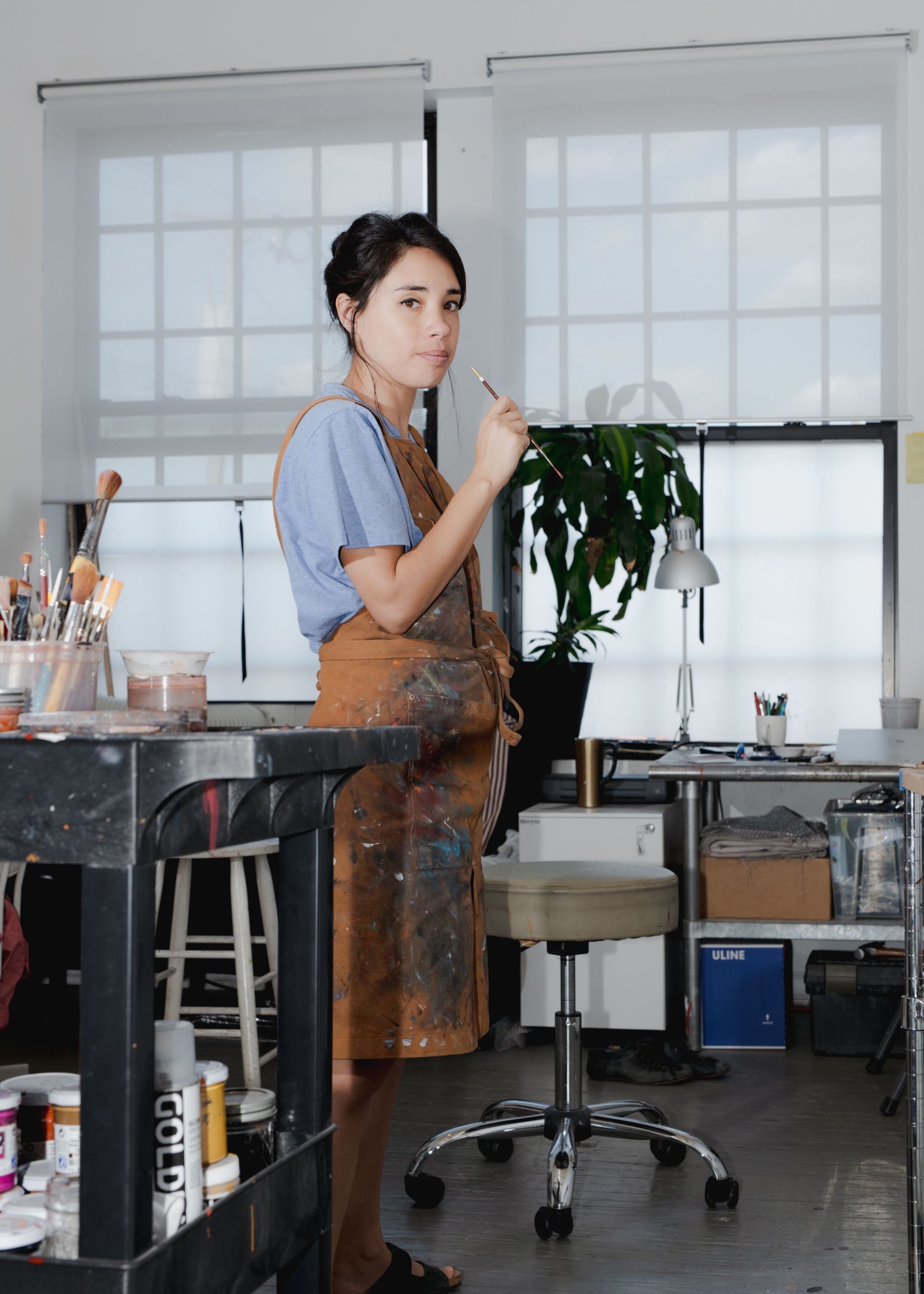 A woman with dark hair swept up behind her head stands in an artist's studio space wearing a paint-speckled apron and holding a think paint brush up in front of her.