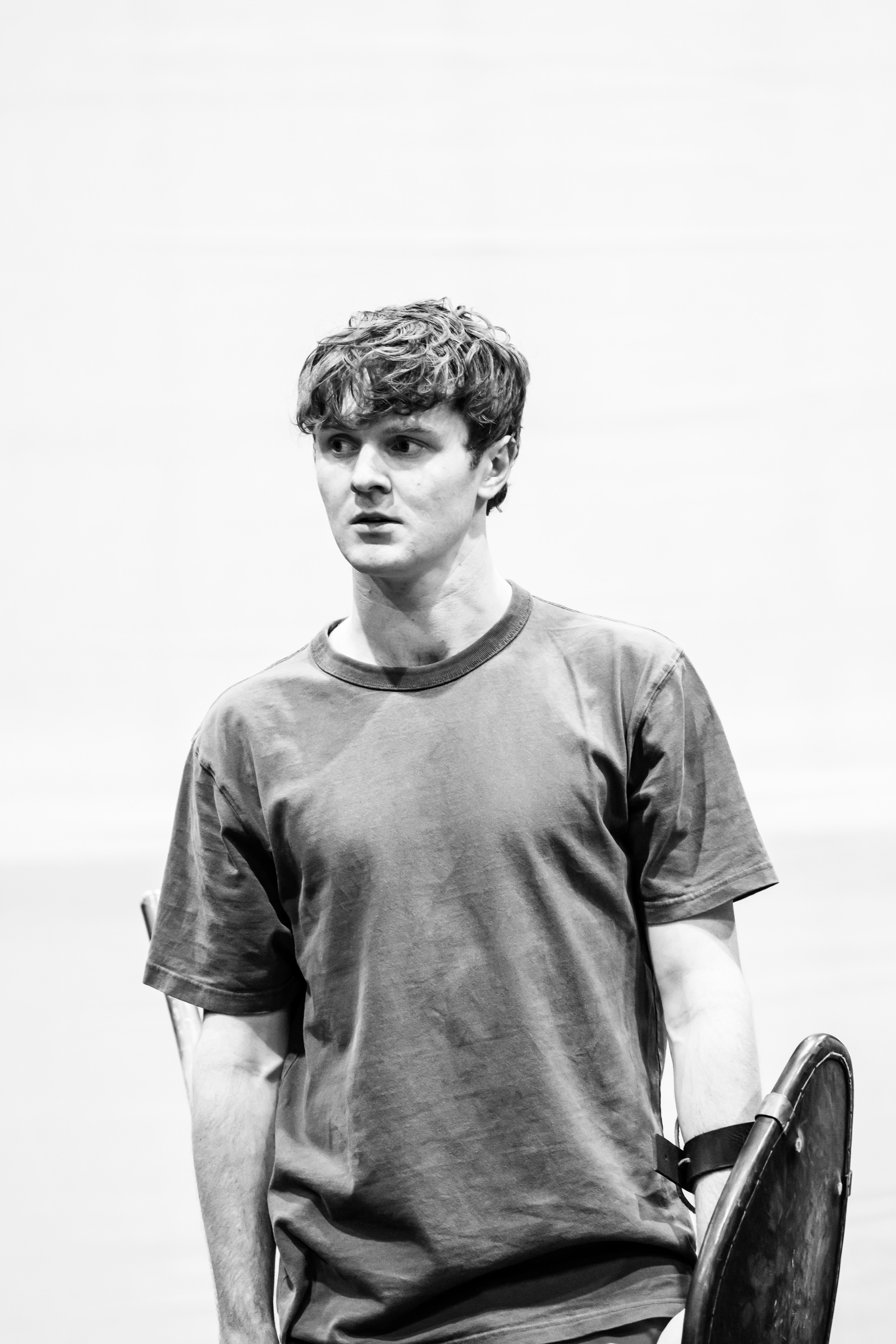 Actor Doug Colling, a white man wearing a gray tshirt, stands with arms to his side in rehearsal. He looks attentively to his side, and hold his arms at his side, his left forearm supporting a prop shield. 