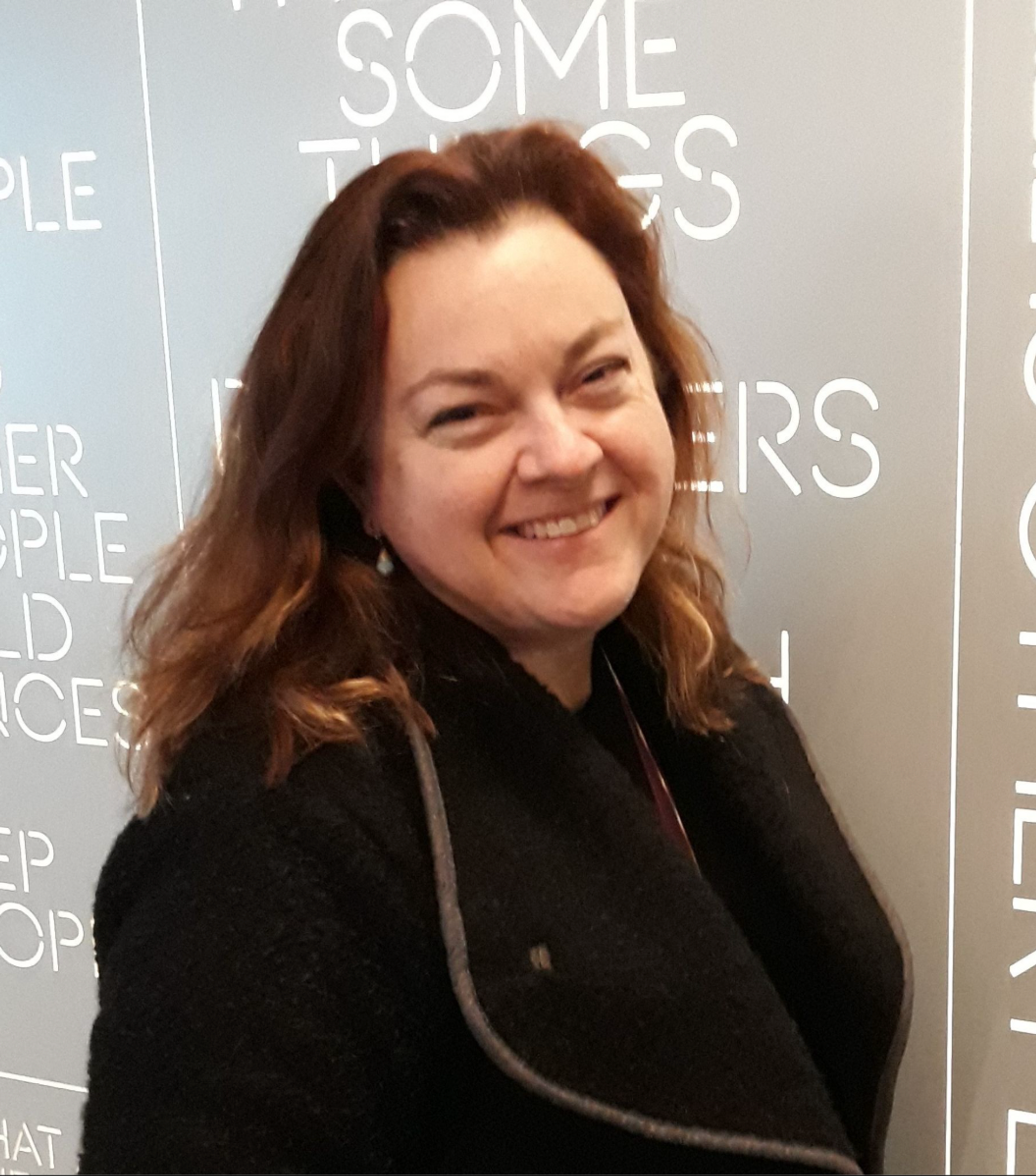 A photo of Deaf actor Pamela Decker-Wright. Pamela is a white woman who has chestnut brown hair parted in the middle that falls to her shoulders. She is seen in three quarters profile, turning slightly toward us, smiling happily. 